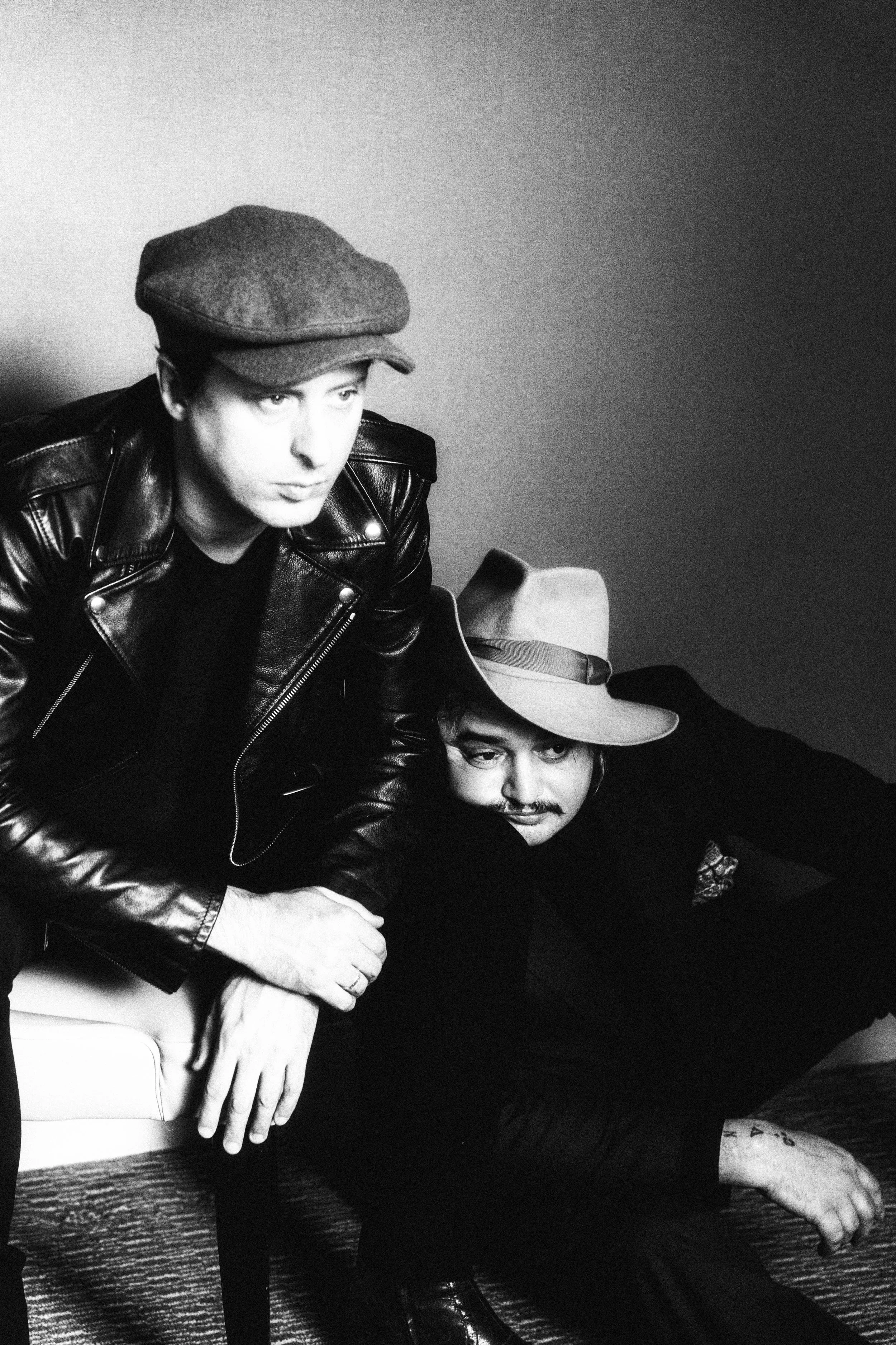Two men in black clothing and hats, one with a leather jacket, are sitting close together against a plain background. One man is resting his arm on the other, who is leaning back with a part of his body visible, showing a tattoo on his leg.