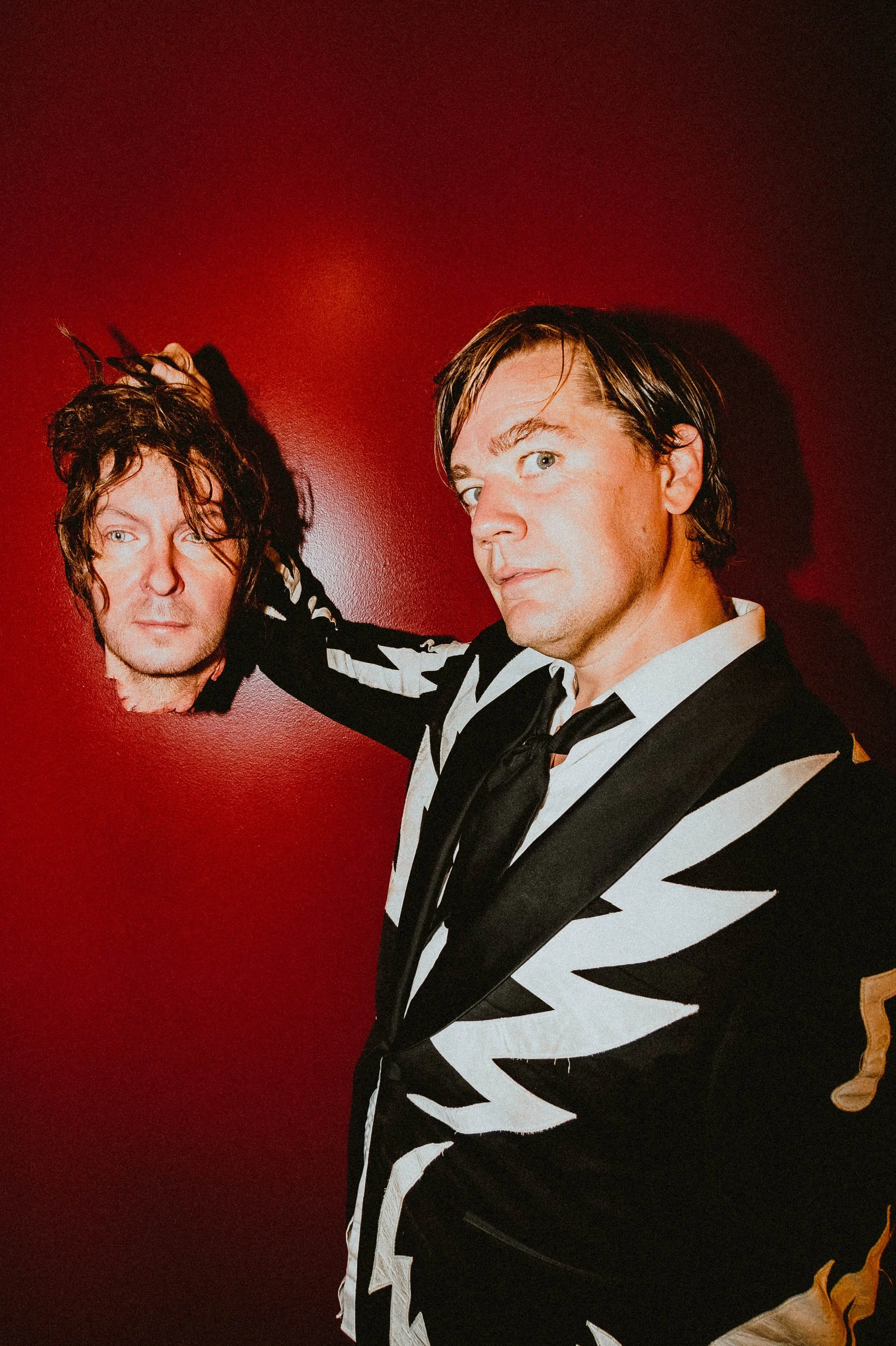 Two men posing against a red wall, one with curly hair and the other with straight hair, wearing black and white patterned outfits.