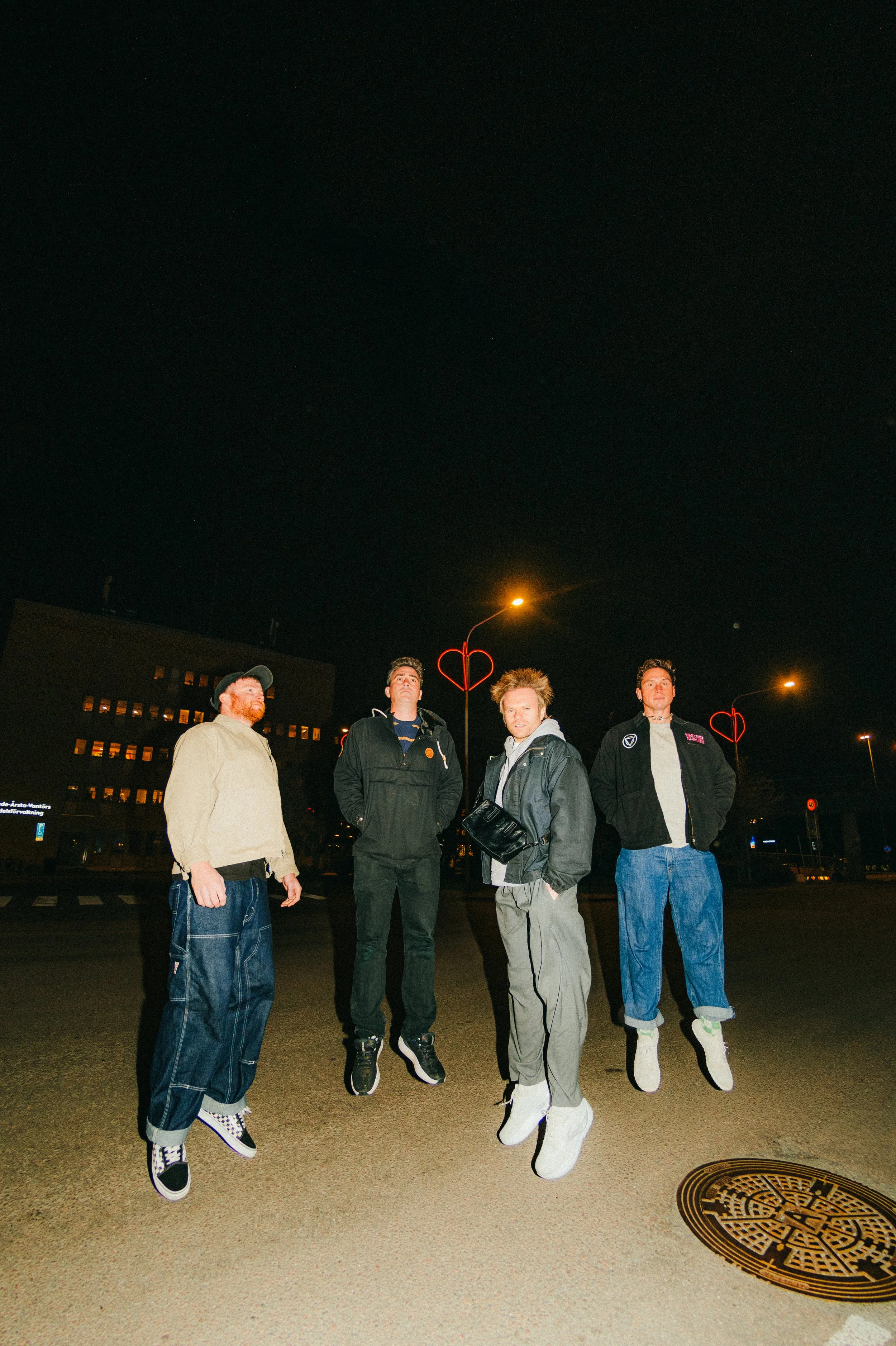 Enter Shikari band men standing on a city street at night, with streetlights and a manhole cover visible in the foreground. The men are wearing casual clothing, including jackets and jeans, and are illuminated by nearby streetlights.