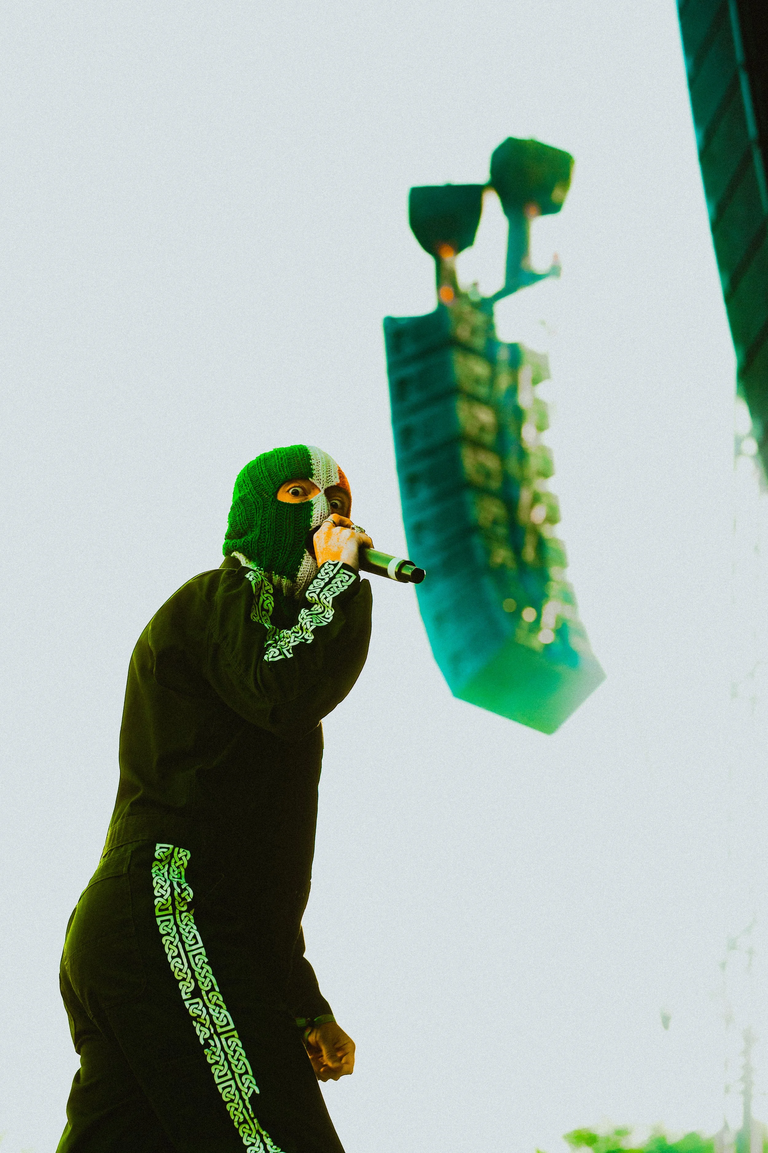 Kneecap singer wearing a black hoodie and a green, white, and black ski mask, speaking into a microphone, with a large green flag or banner nearby.