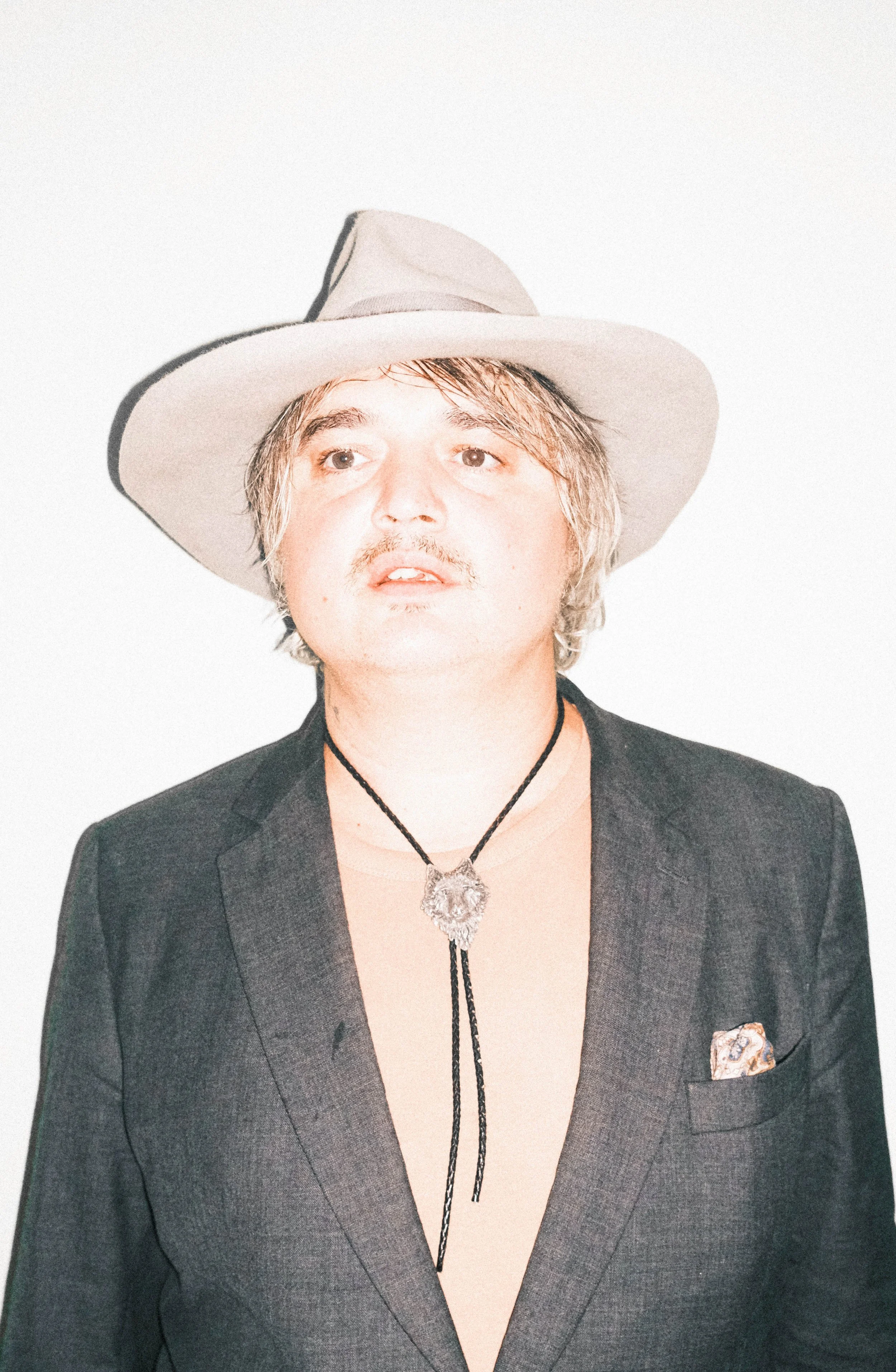 Pete Doherty wearing a wide-brimmed hat, black blazer, and a necklace with a lion pendant, posing against a plain white background.