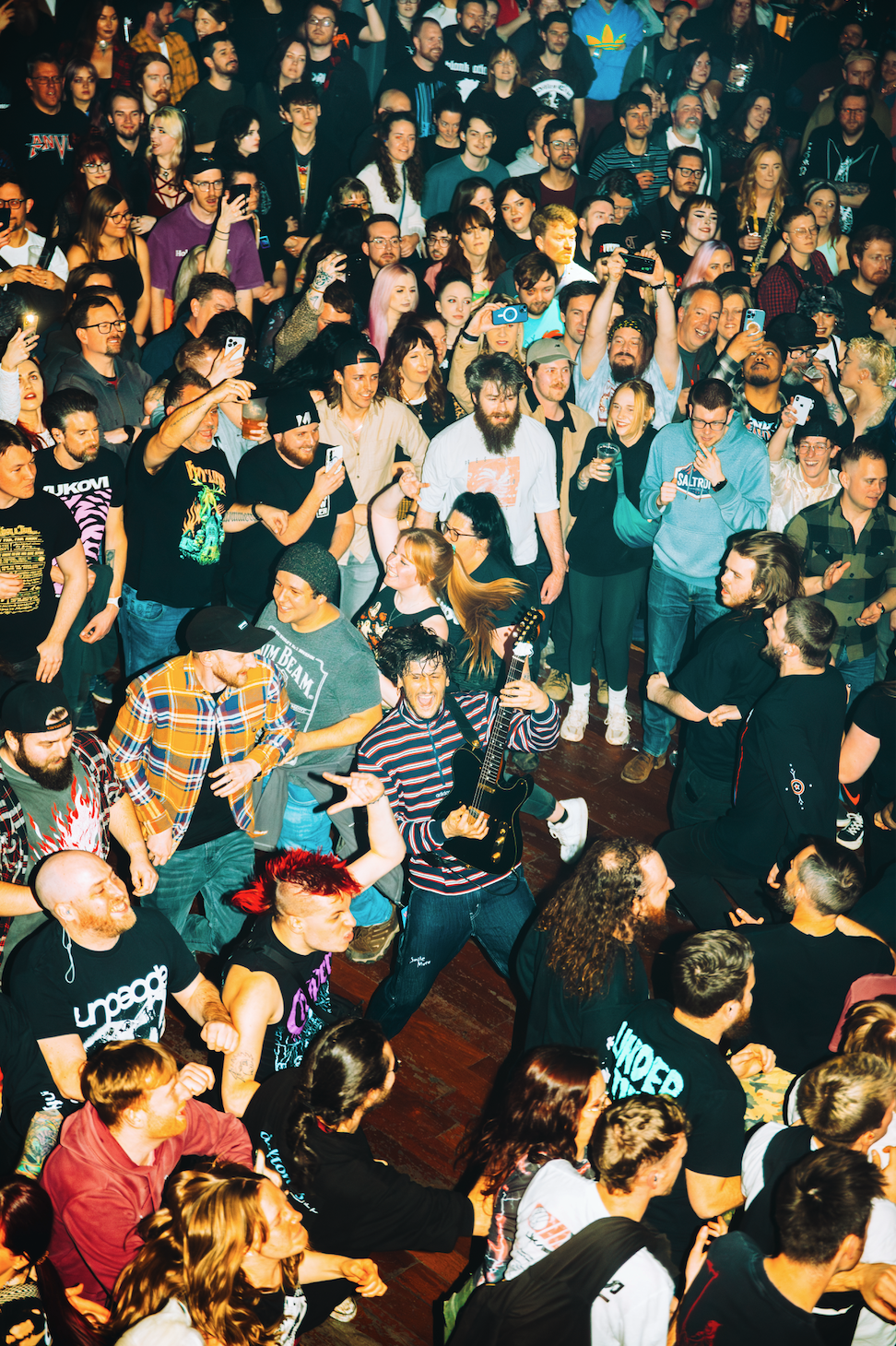 A crowd of people at a concert, with some taking photos or videos, a guitarist appears to be performing, and the audience is lively and engaged.