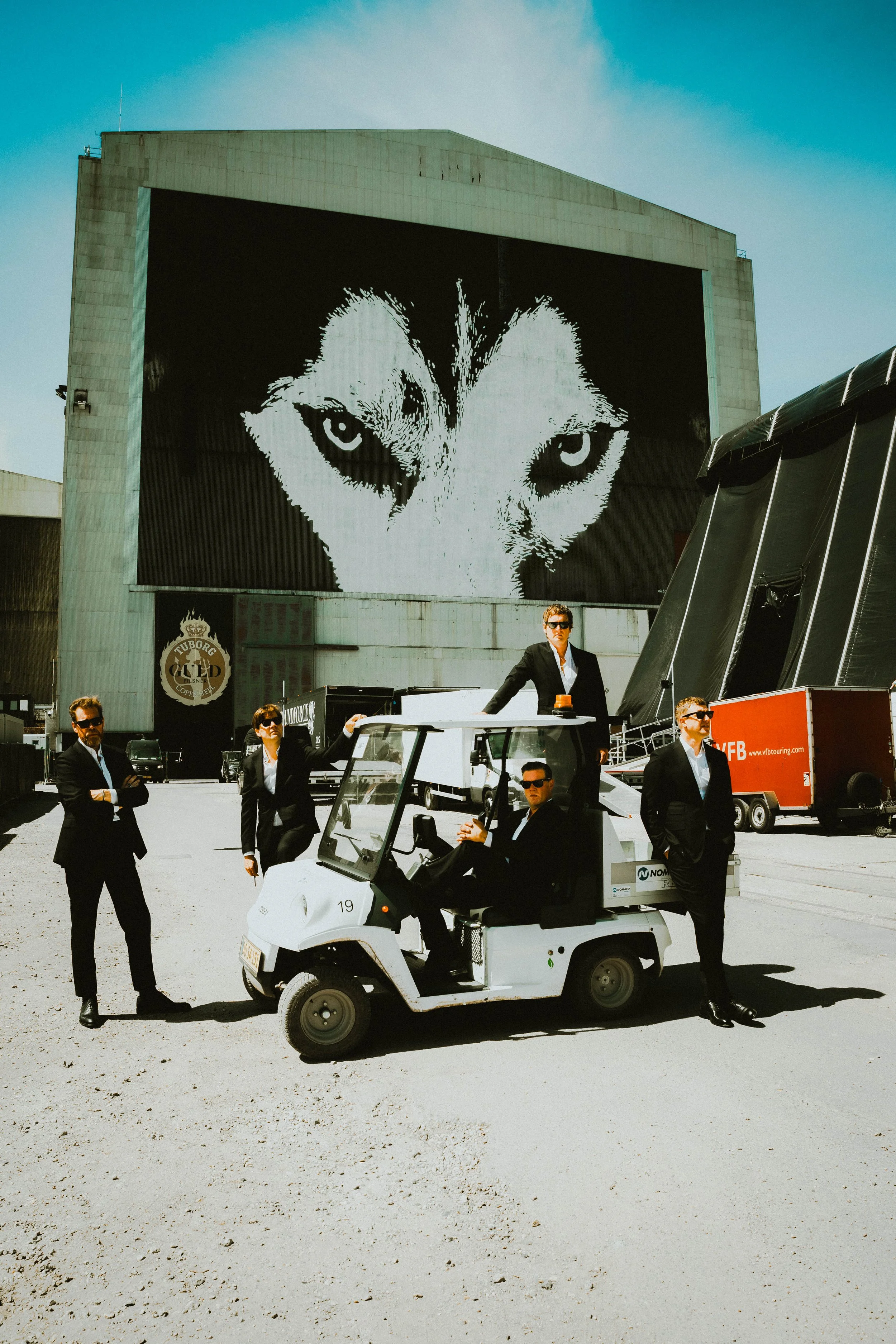 The Hives band in black suits and sunglasses standing and sitting around a small white golf cart in an outdoor area. In the background, a large building displays a prominent black-and-white image of a cat with piercing blue eyes.