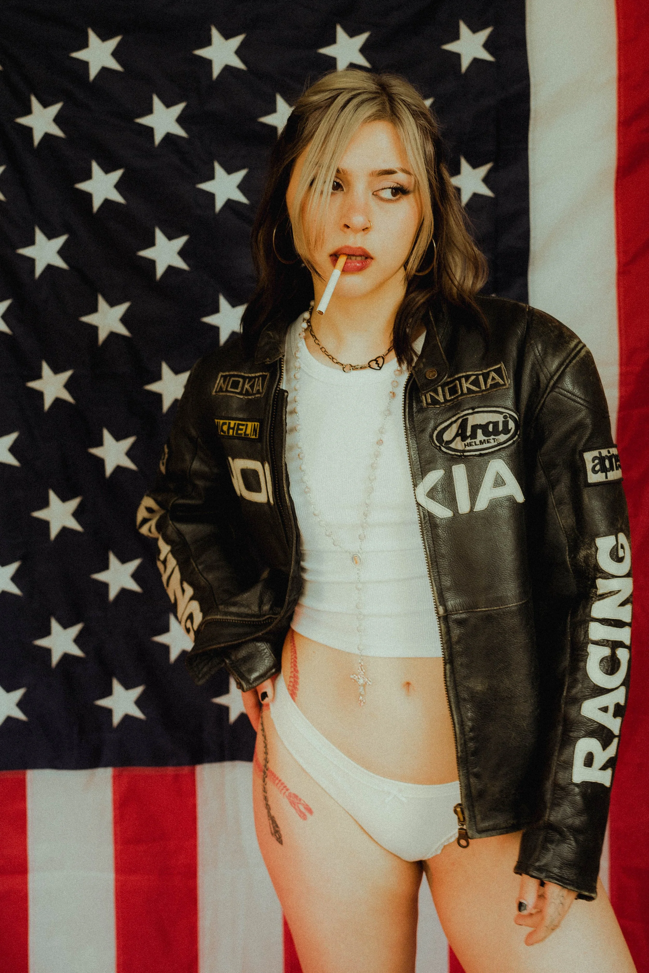 A young woman with shoulder-length dark hair and a blond streak, wearing a black leather jacket with various patches, a white shirt, and white underwear, stands in front of an American flag backdrop. She has a cigarette in her mouth and a piercing on
