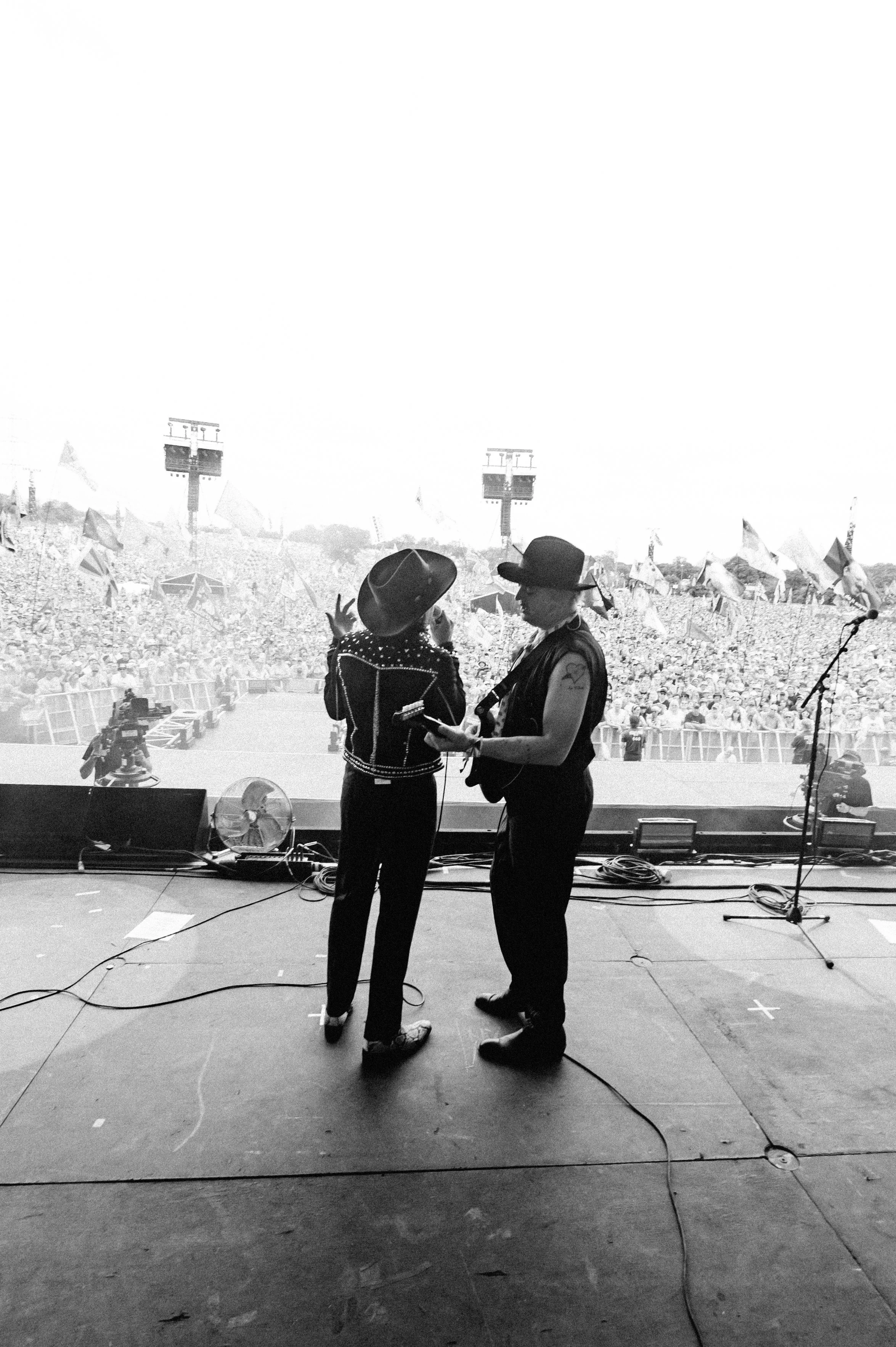 Pete Doherty and Carl Barat from The Libertines wearing dark outfits and hats stand on stage in front of a large crowd at Glastonbury, with flags and banners visible in the background.