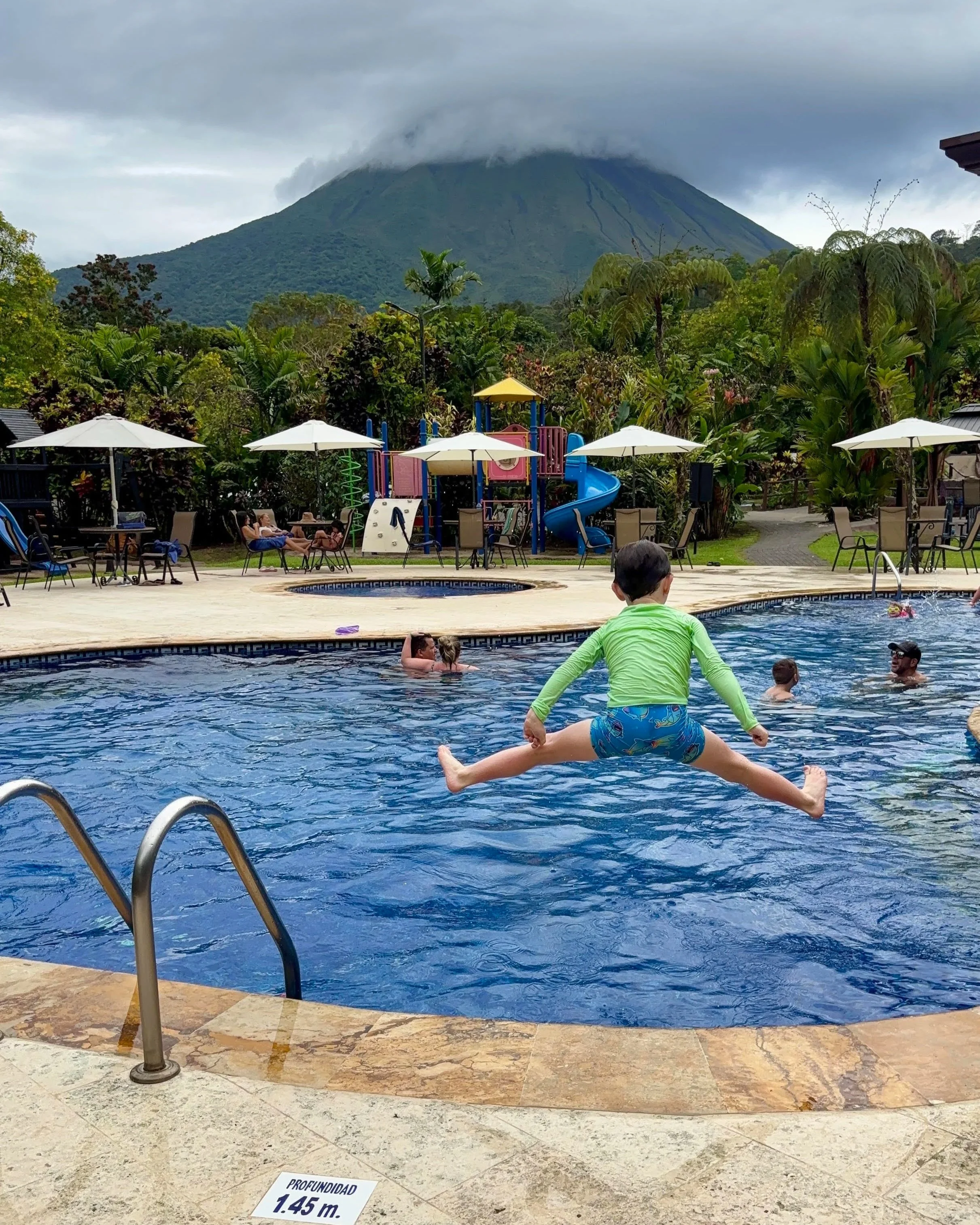 Swimming pool with children playing, surrounded by chairs and umbrellas, with lush green trees and a mountain with clouds in the background.