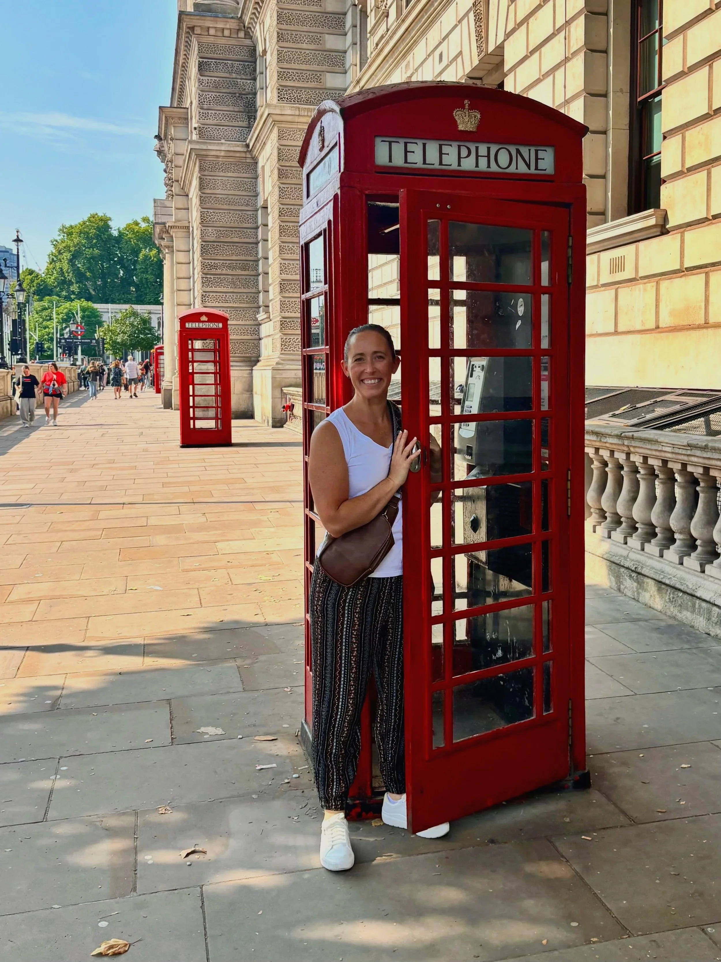 A woman smiling and standing inside a traditional red British telephone booth on a city sidewalk, with a building and other telephone booths visible in the background.