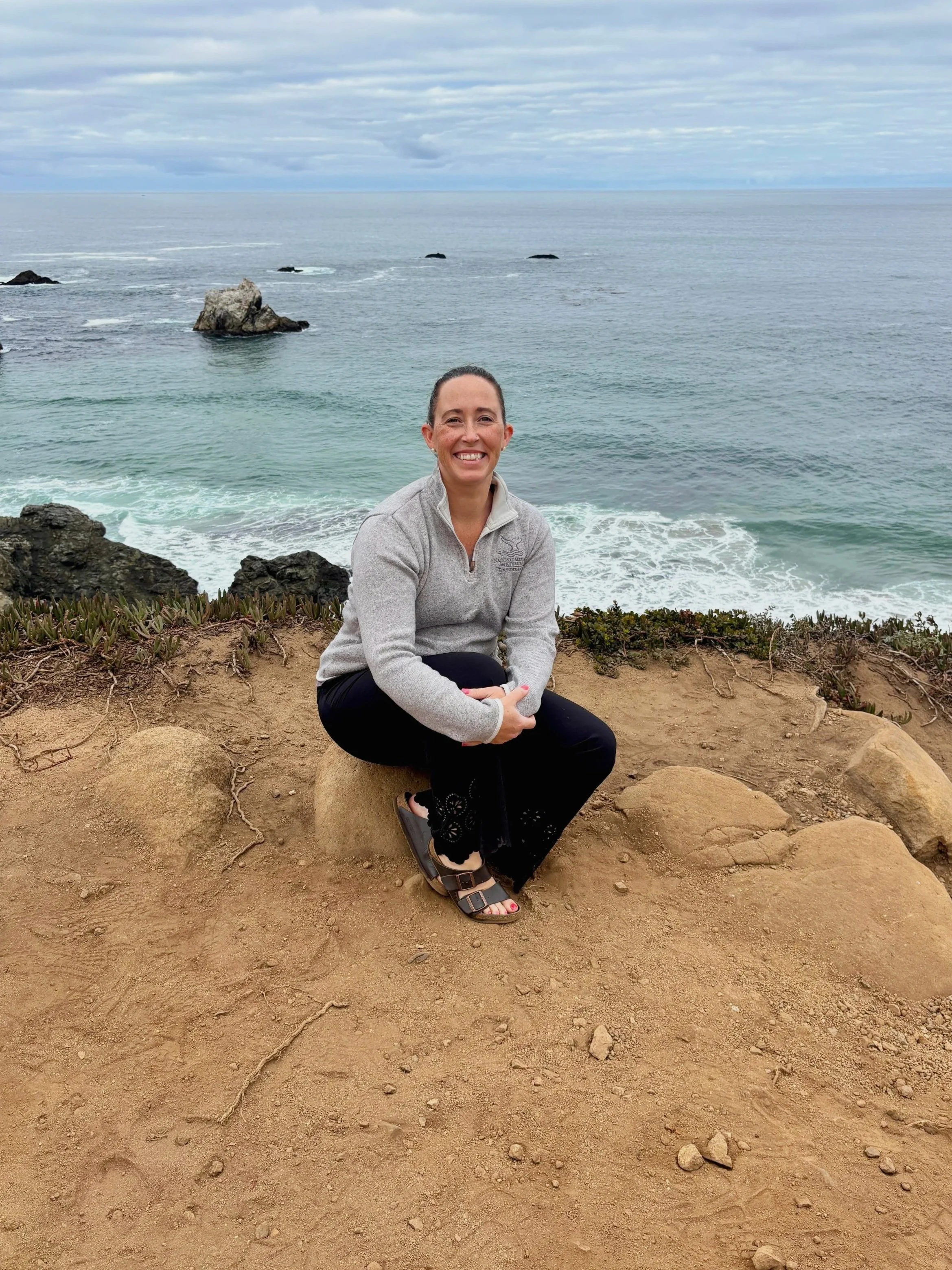 A woman sitting on a large rock by the coast, smiling, with ocean waves and rocks in the background, under cloudy sky.
