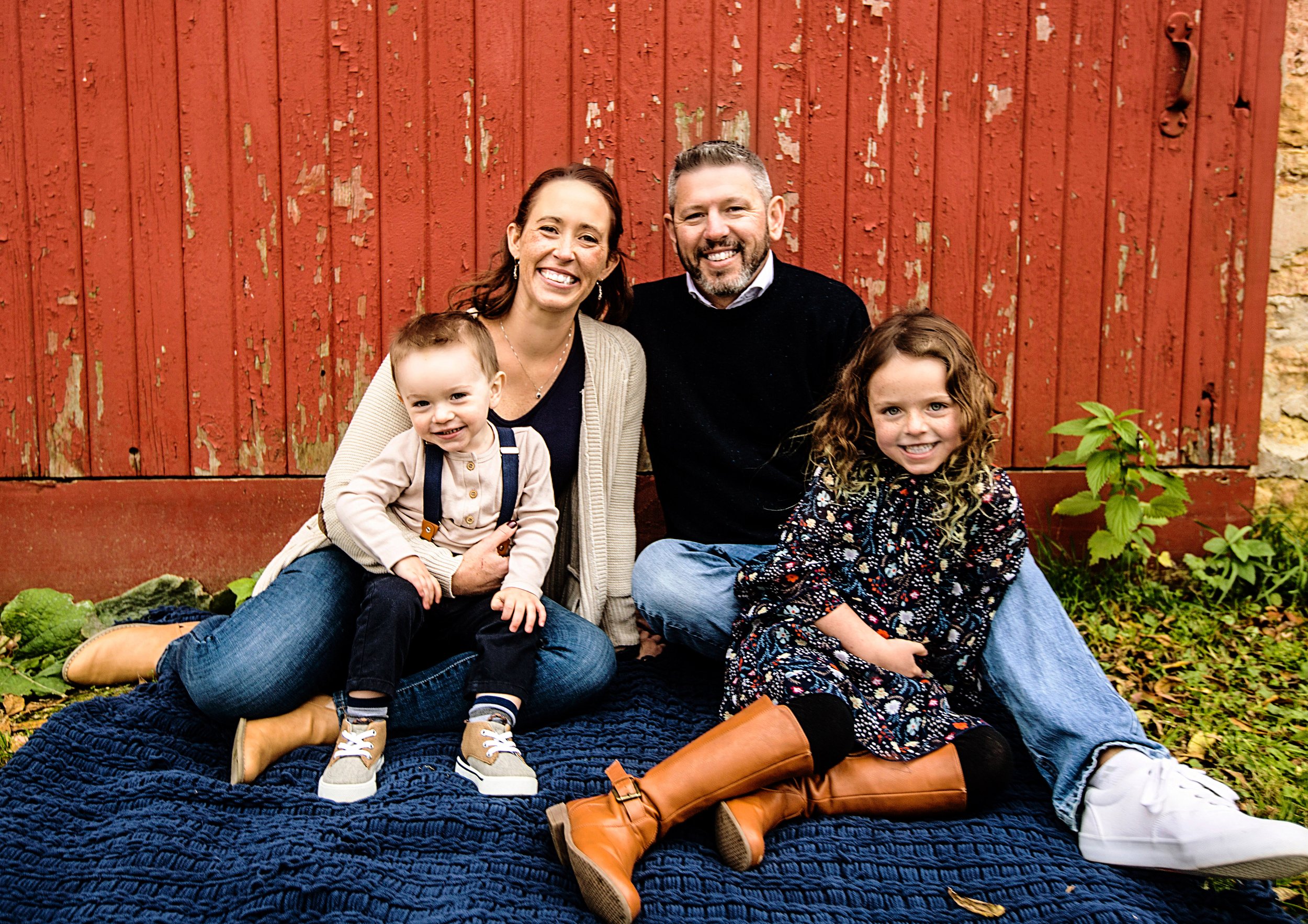 A family of four, including two children and their parents, smiling and sitting outdoors in front of a weathered red wooden barn. The family appears happy, with the children sitting on their parents' laps. The ground is covered with green plants and leaves.