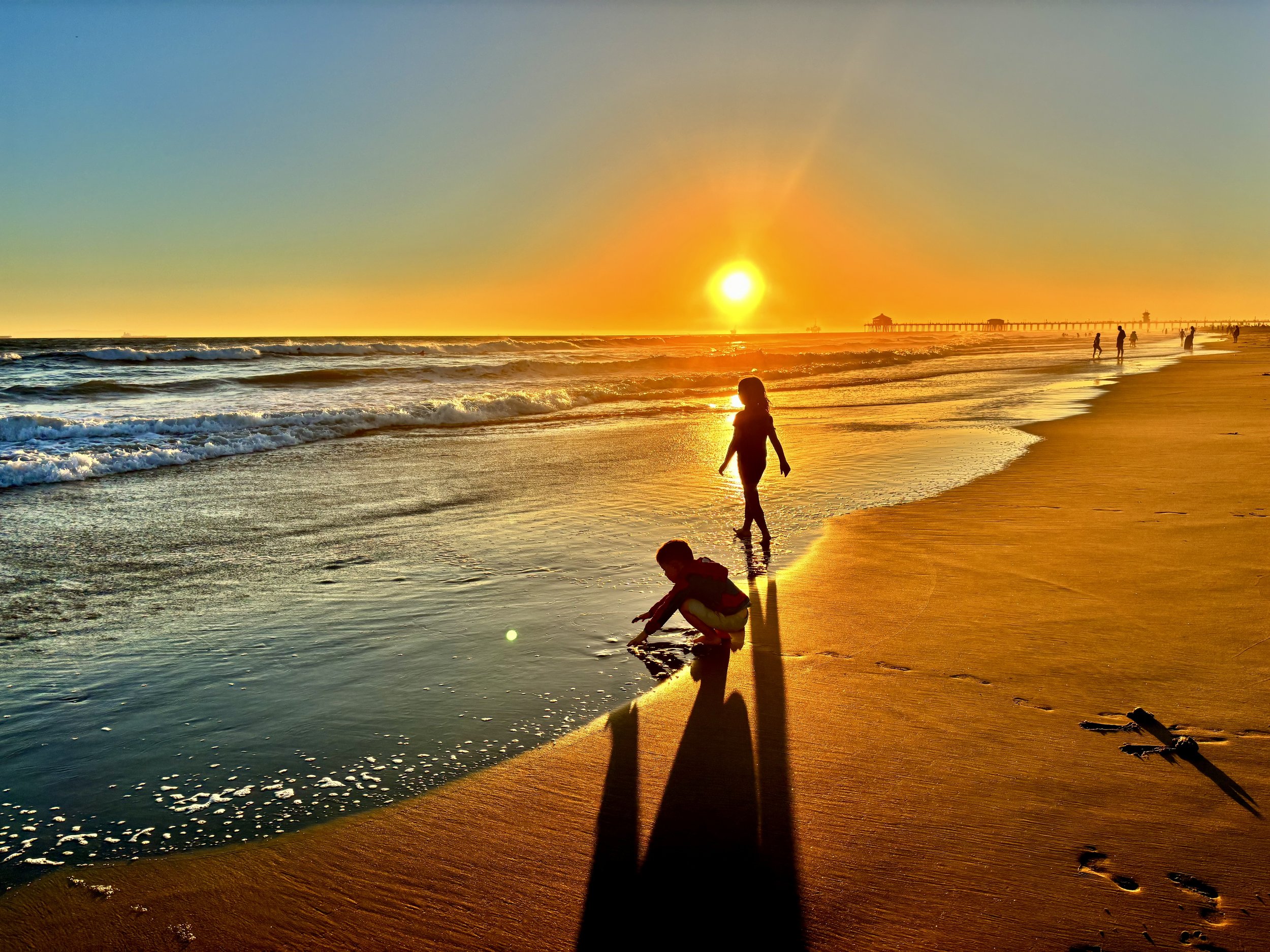 Children playing and walking on the beach during a sunset with a pier in the background.