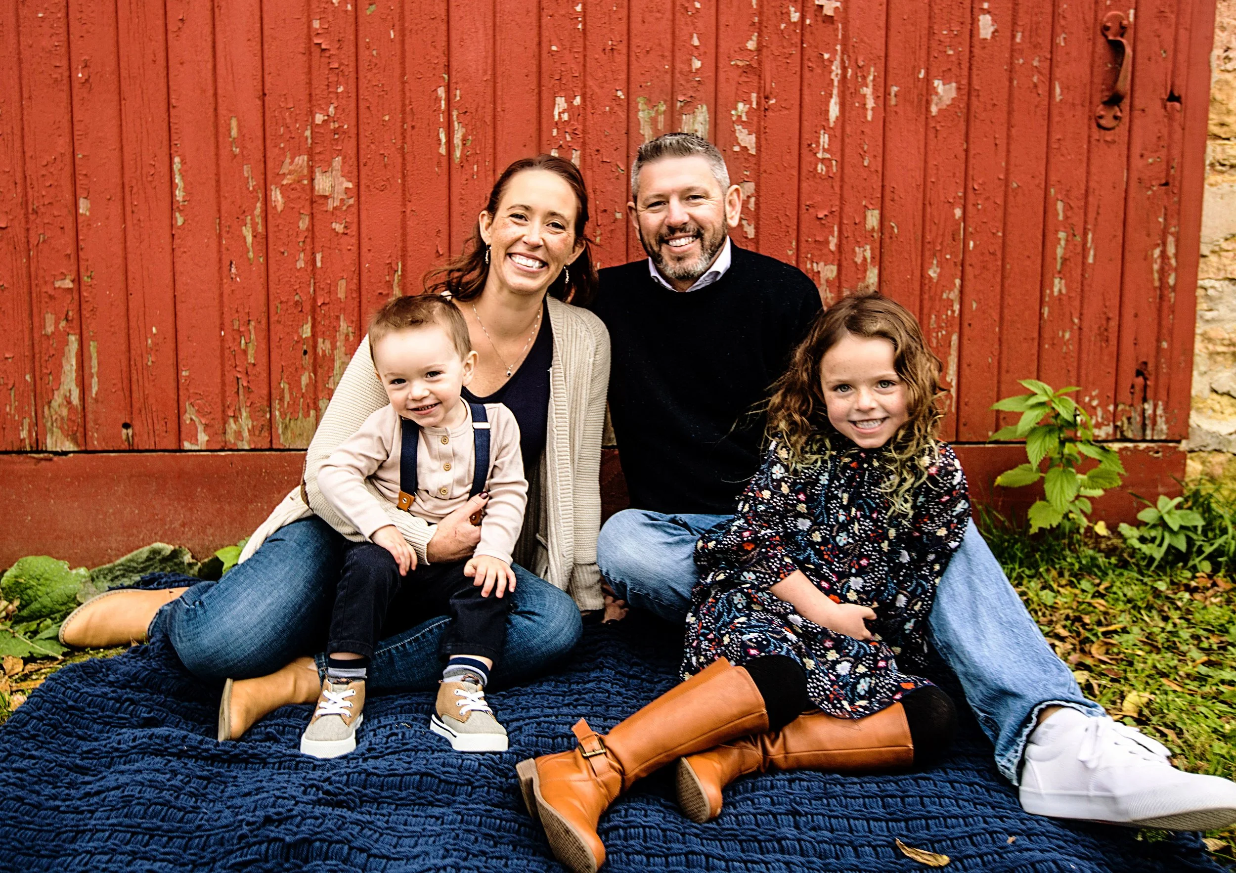 A family of five, including two children and two adults, sitting and kneeling on a dark blue quilt in front of a red wooden barn, smiling at the camera during daytime.