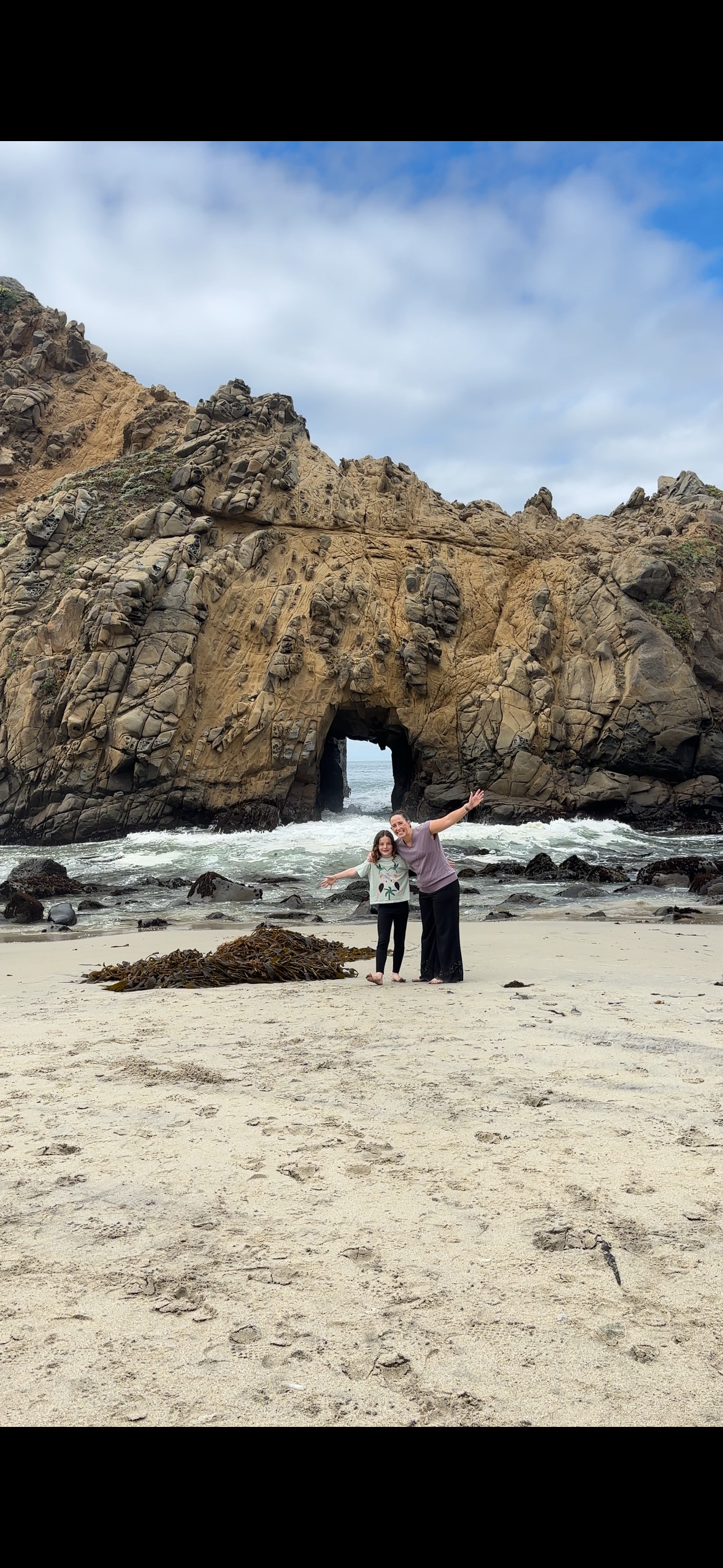 A woman and a girl stand on a sandy beach with their arms wide open, smiling. Behind them is a large rock formation with a natural archway and smaller rocks, ocean waves, and a cloudy sky.