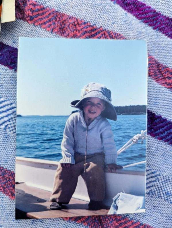 A smiling young child standing on a boat deck next to water, wearing a large sun hat, a light sweater, and brown pants, with a scenic lake and forest in the background.