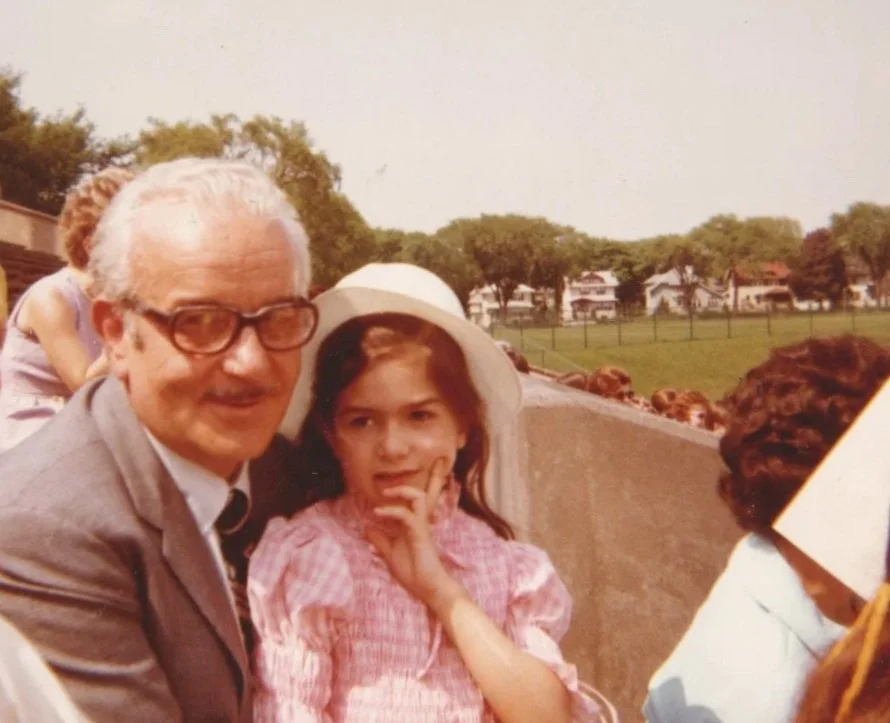 John H. Secondari and his daughter, Linda, at the graduation of his son JG Secondari from college. They are posing outdoors during daytime, with trees and houses in the background; the girl is wearing a wide-brimmed hat and a pink checkered dress.