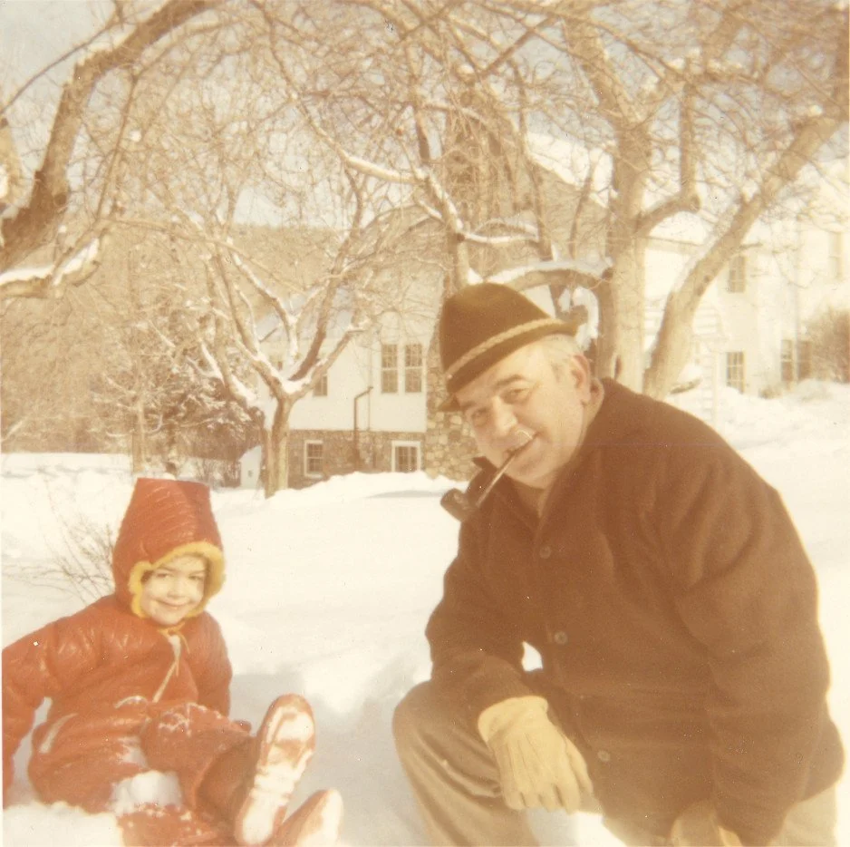 John H. Secondari and his daughter Linda Secondari are sitting in the snow during winter in Monterey, Massachusetts. He is wearing a dark coat, hat, and gloves, with a pipe in his mouth. she is dressed in a red snowsuit with a hood and is smiling.