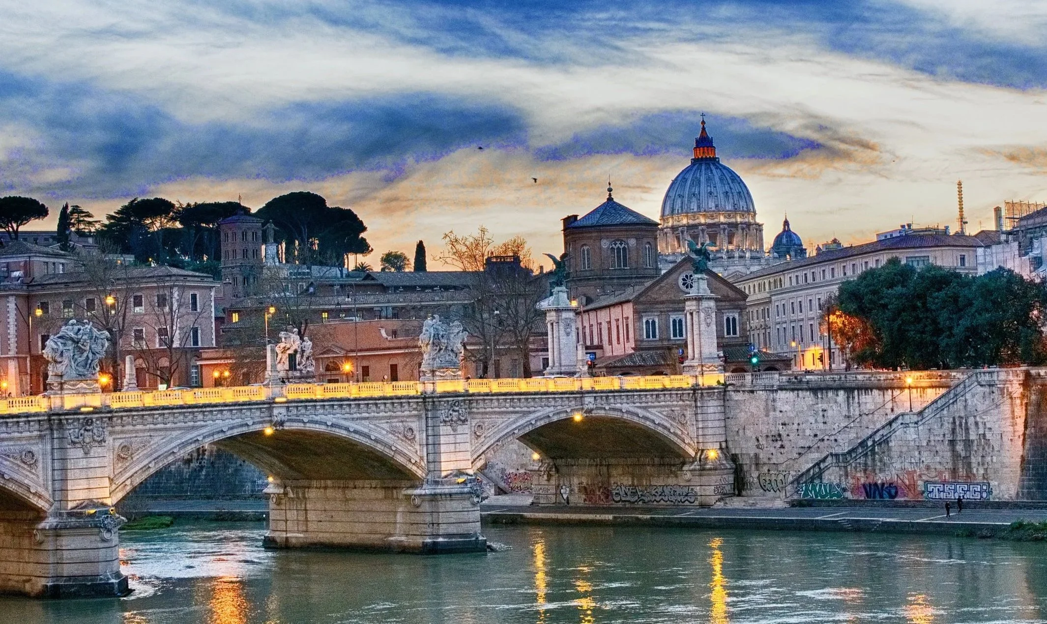 Sunset over Rome with St. Peter's Basilica and the Bridge of Angels crossing the Tiber River, illuminated statues and buildings.