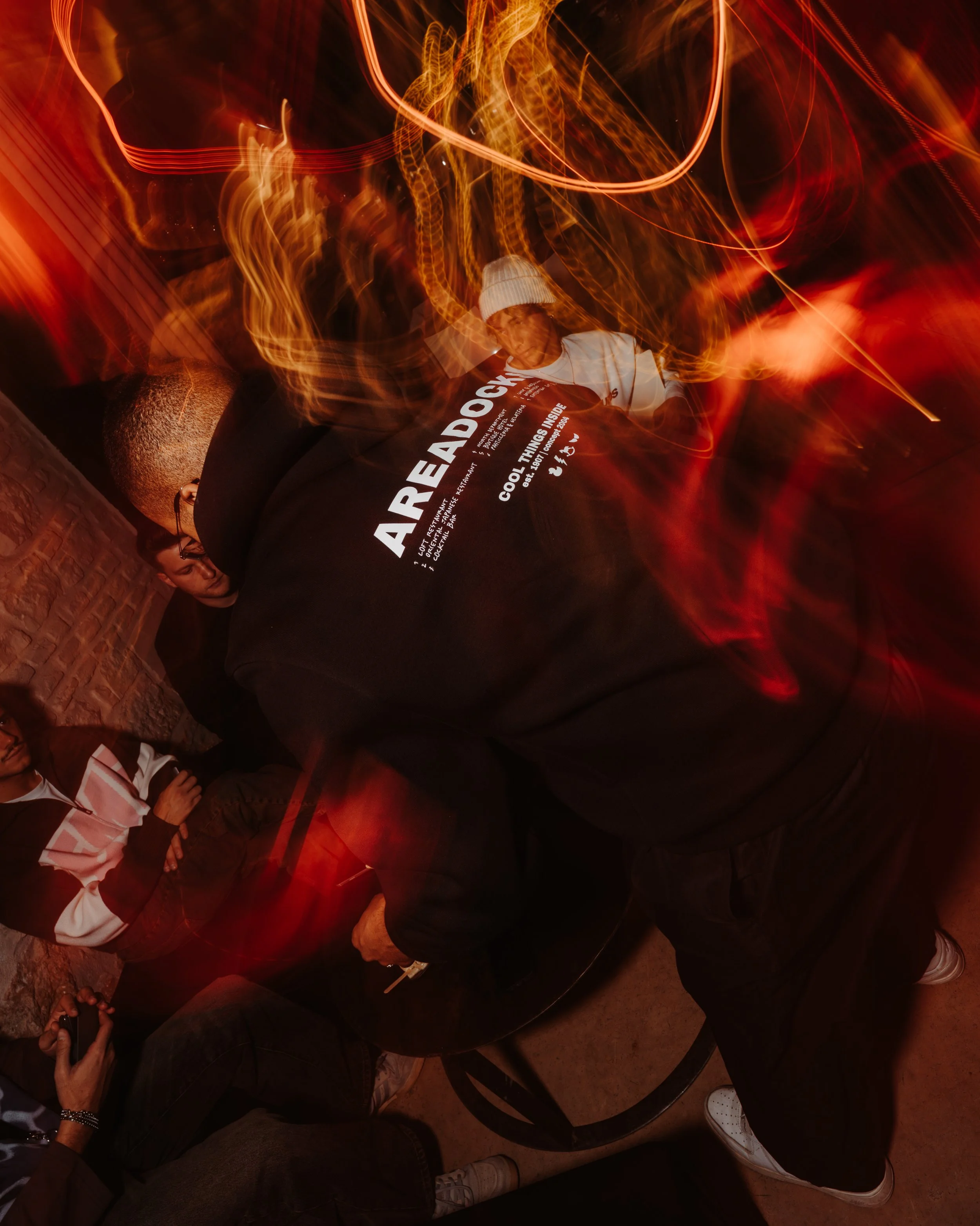 Person wearing a white beanie and white shirt sitting on a black chair, with swirling red and orange lights in the background, creating a dynamic, blurry effect.