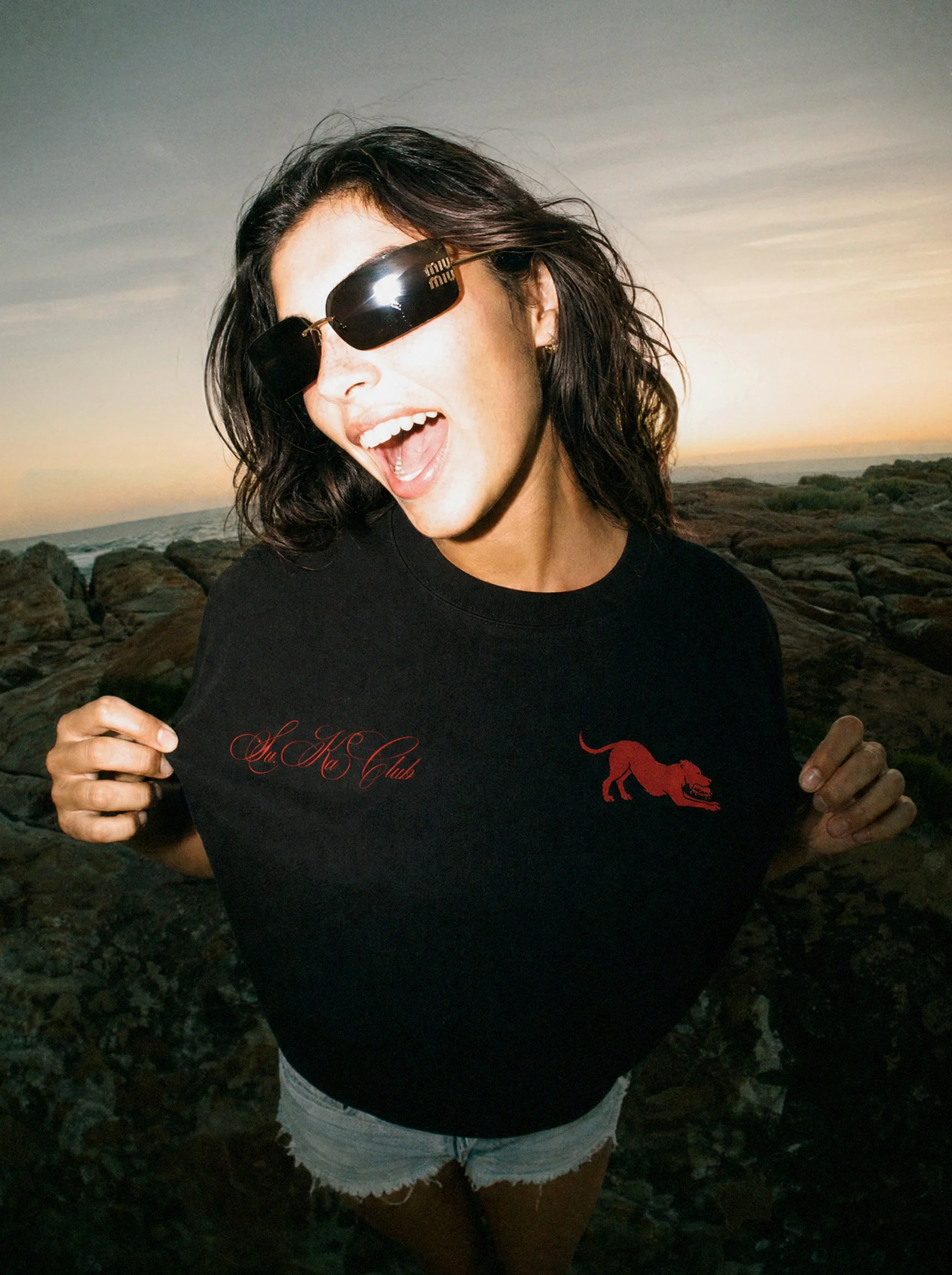 A woman at the beach during sunset, wearing sunglasses and a black Su.Ka Club T-shirt with a red lion graphic and red text, and denim shorts, smiling with her mouth open, with rocks and the ocean in the background.