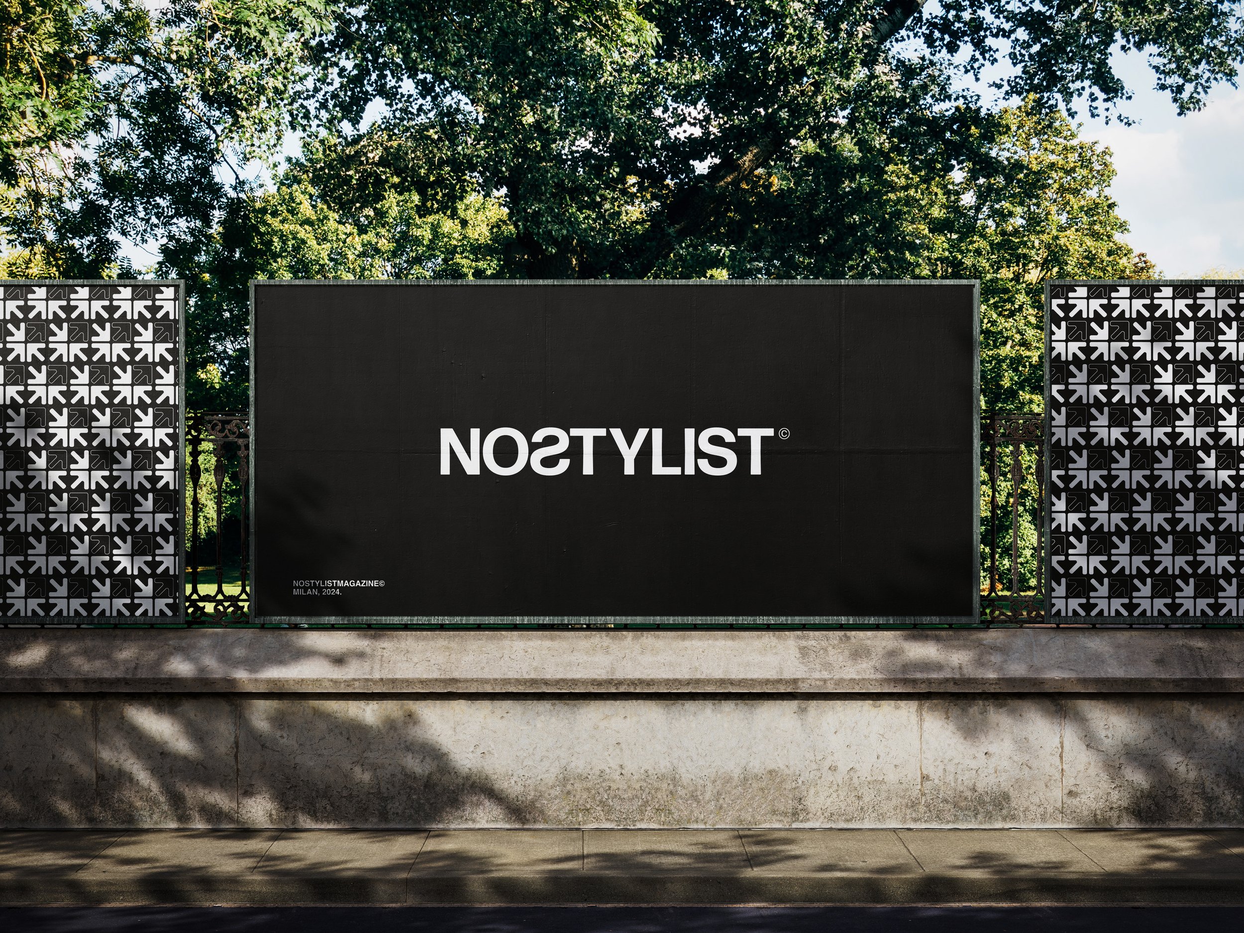 A large black billboard with white text reading 'NOSTYLST,' flanked by decorated black and white patterned panels, set outdoors with trees and blue sky in the background.