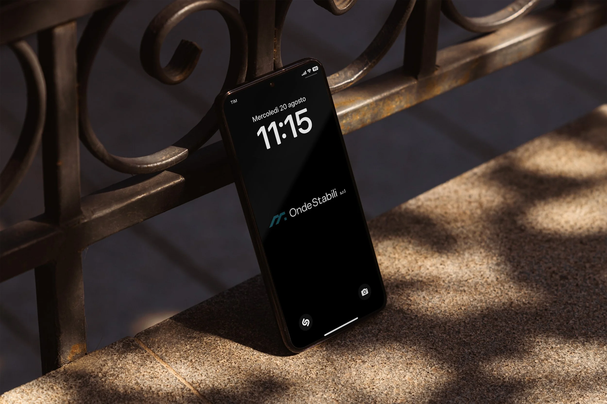 A smartphone resting on a rustic, metal outdoor table with a black surface, displaying the time 11:15 and the date August 20, Wednesday. The phone is partially propped against the edge of the table, with the screen showing app icons, including a camera and a messaging app. The background includes a stone surface and a shadow cast by the table.