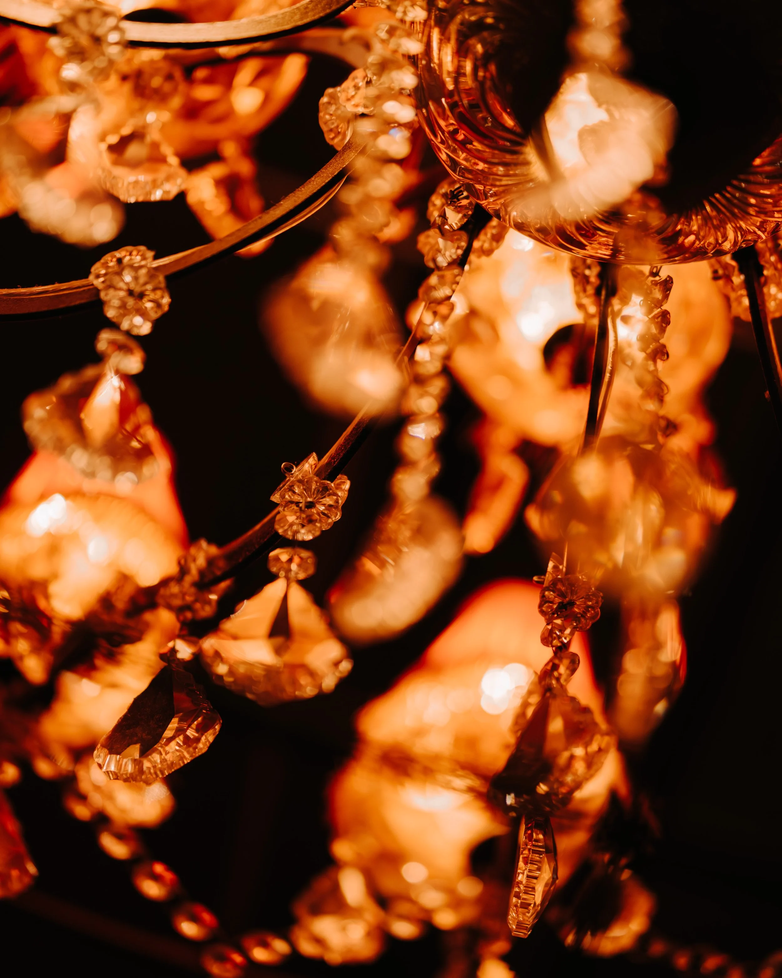 Close-up of an ornate, glowing chandelier with intricate hanging crystals and metallic details, emitting a warm, amber light.