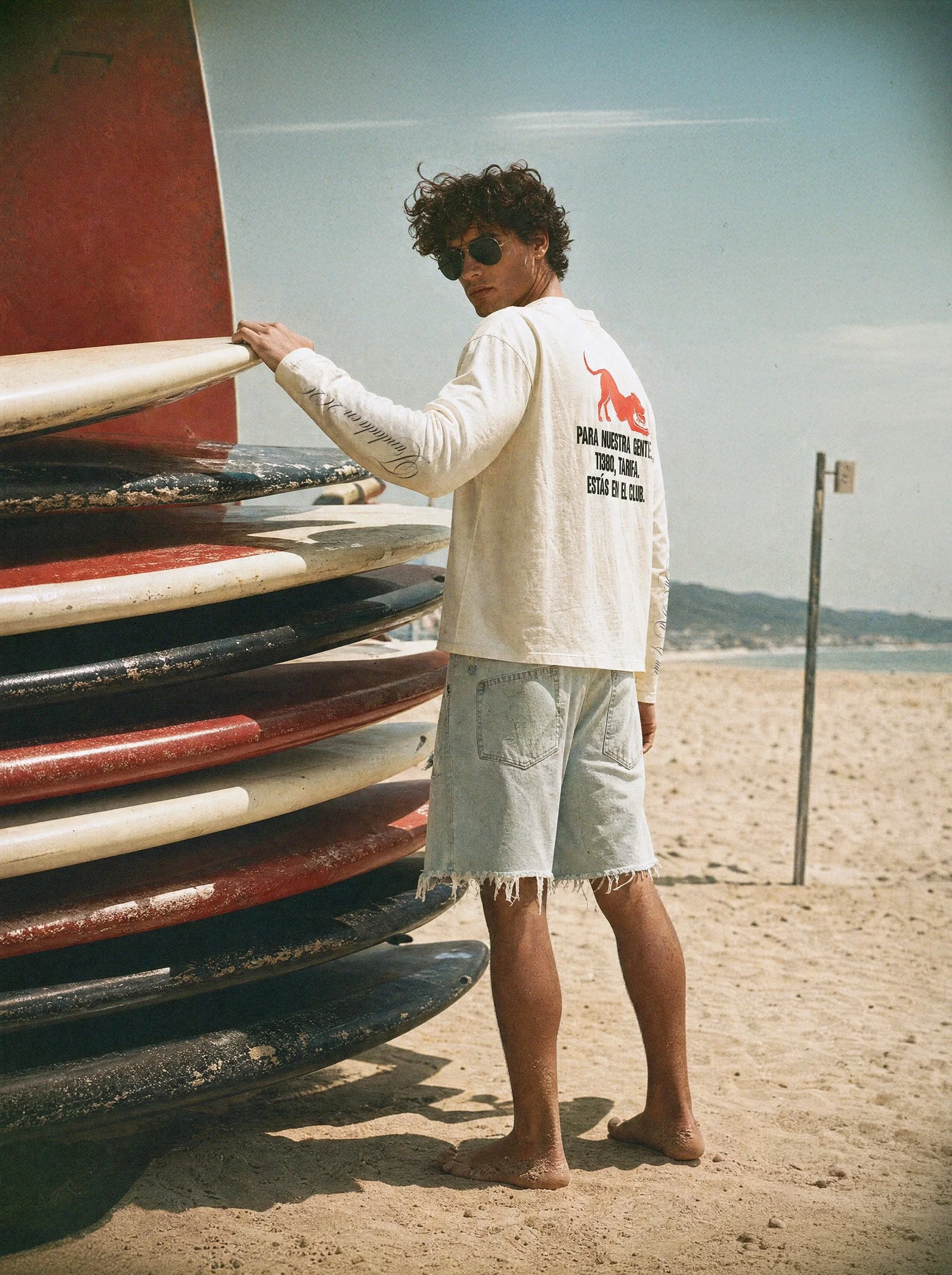 A young man with curly hair and sunglasses standing on the beach, holding onto a stack of surfboards.