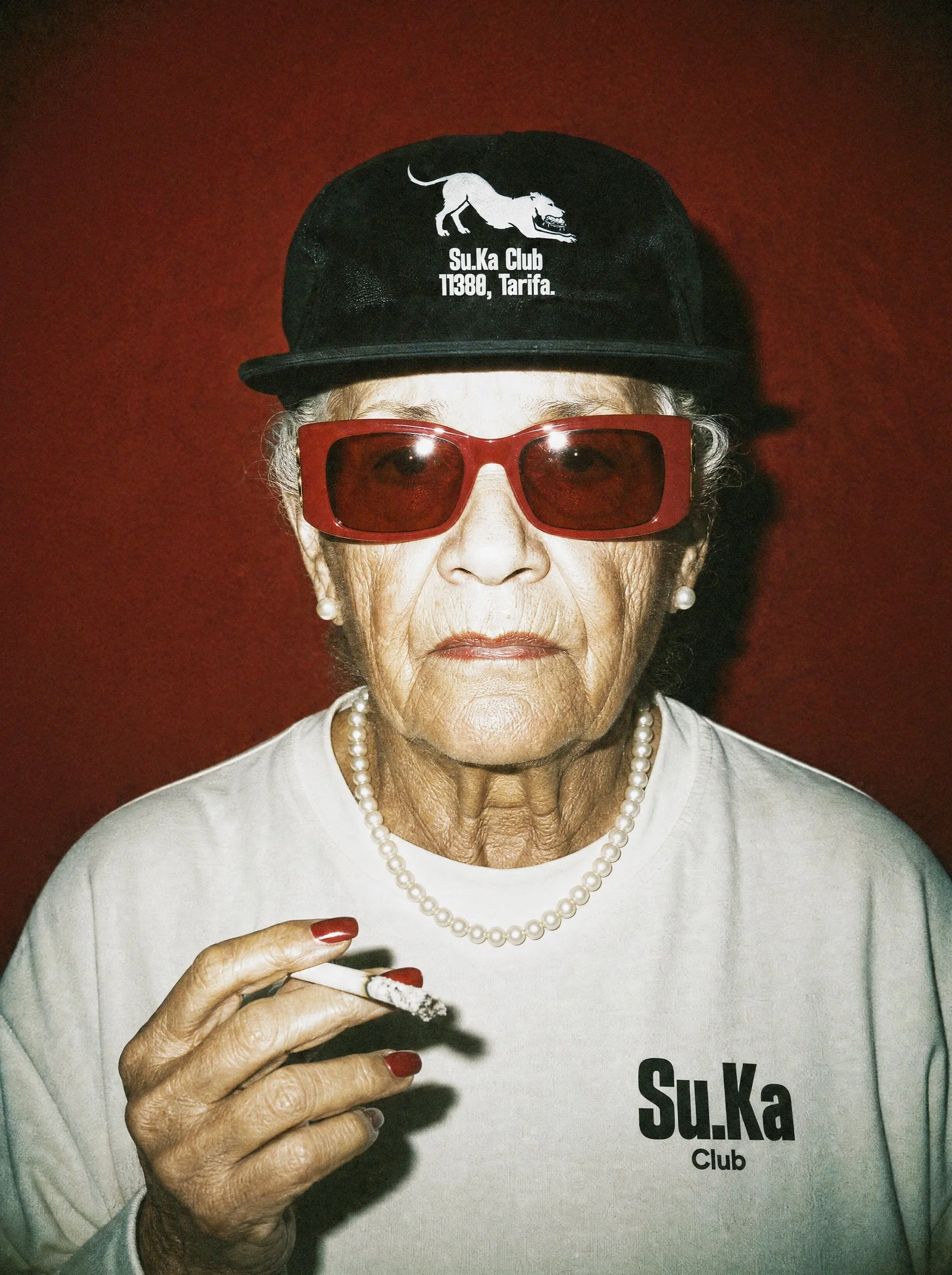 Close-up of an elderly woman with gray hair, wearing large red sunglasses, pearl necklace, and a light-colored shirt with text reading 'Su.Ka Club'. She is smoking a cigarette.