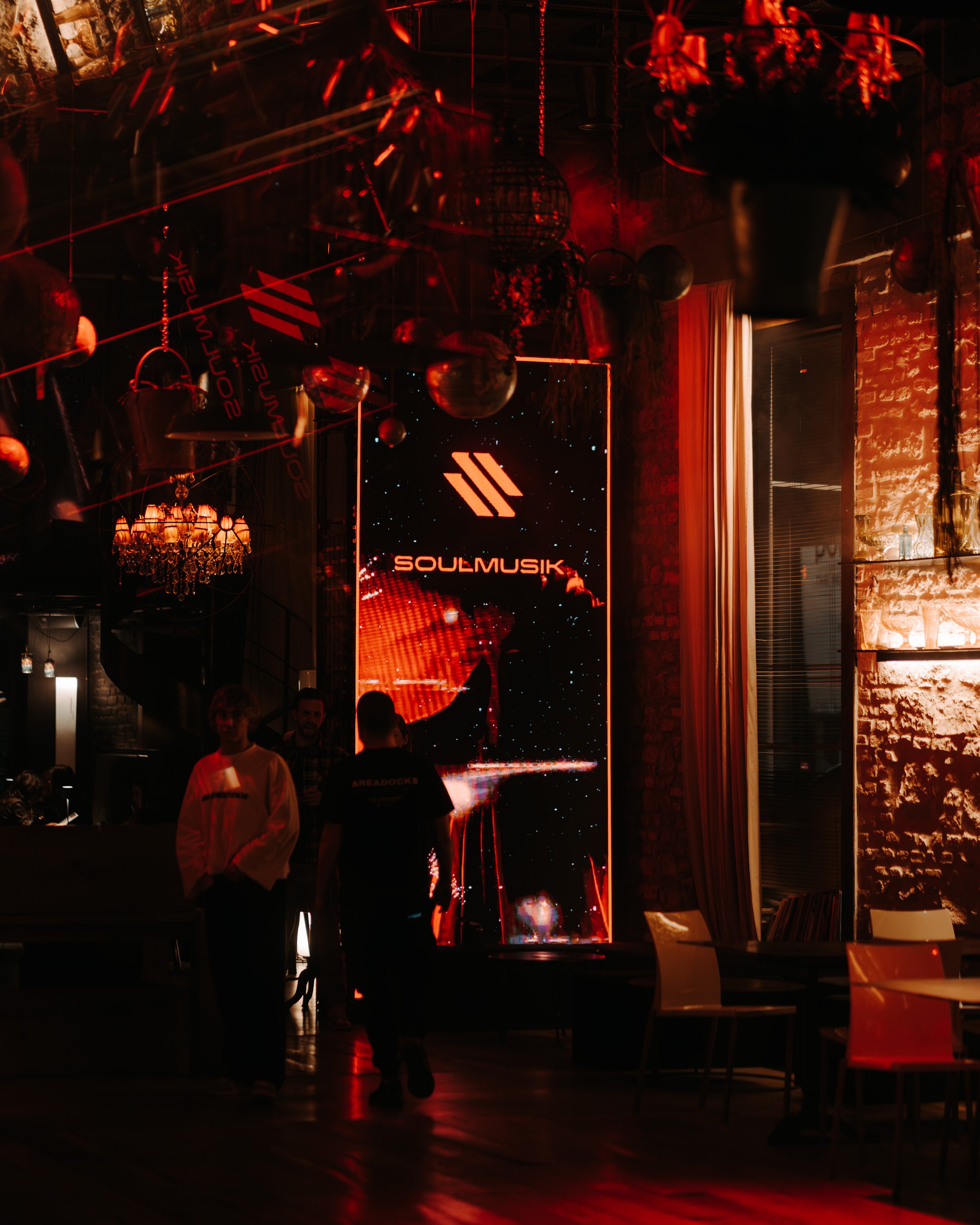 Dimly lit bar or lounge with red lighting, hanging spherical decorations, and a large digital screen displaying the logo 'SOULMUSIK.' There are people standing and sitting, with some silhouettes visible against the lighting.