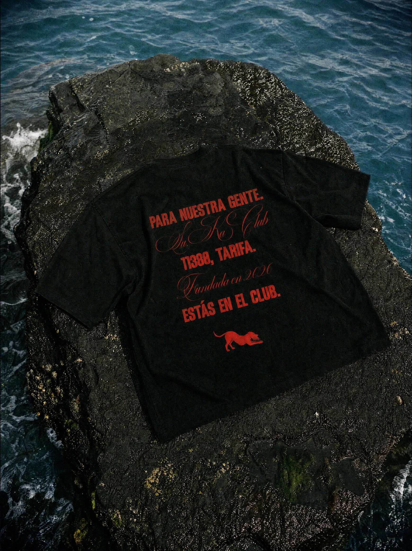 A black Su.Ka Club T-shirt with red and gold lettering, resting on a wet rock near the ocean.