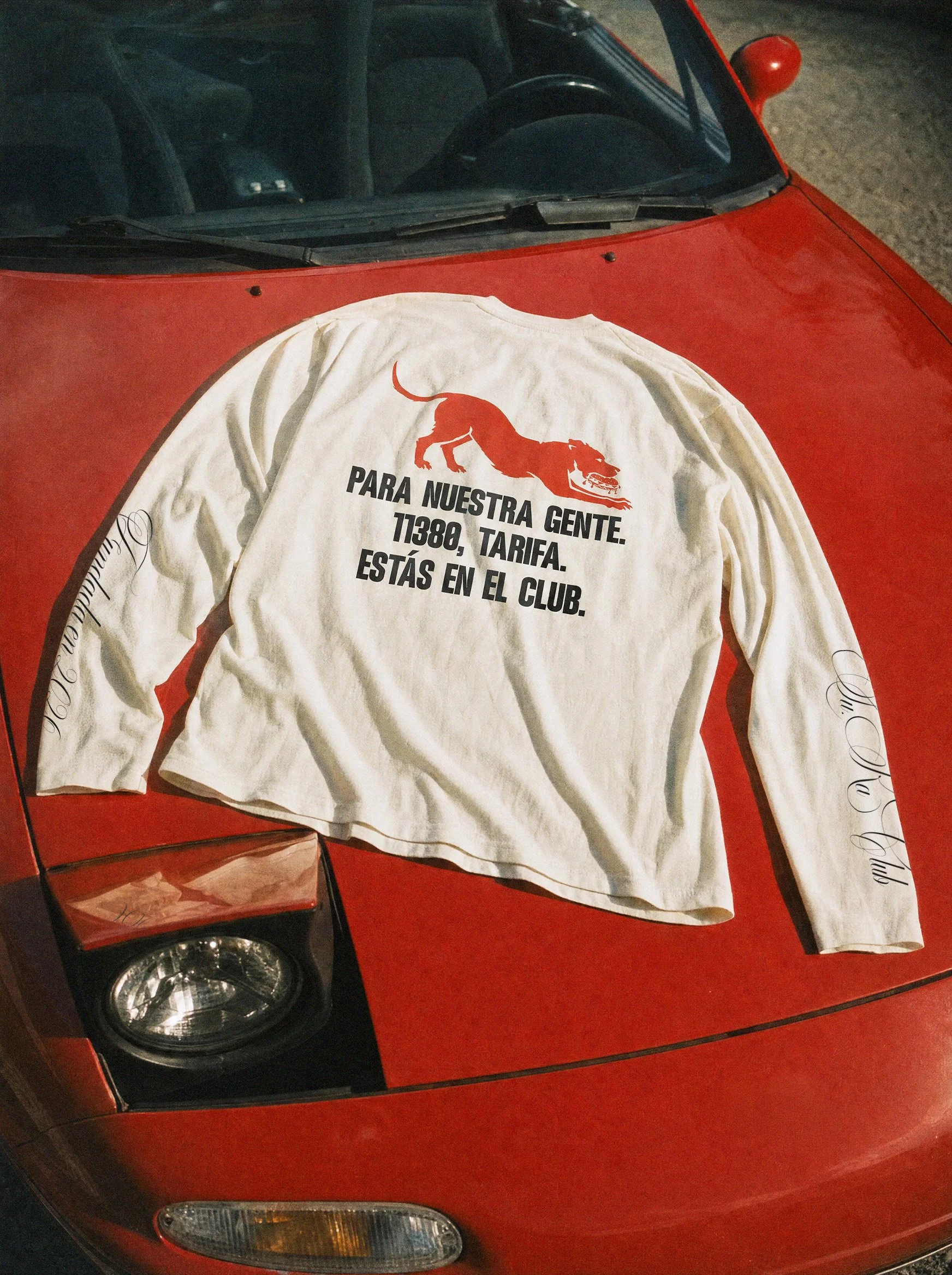 A white T-shirt with a red graphic of a dog and the text in Spanish 'Para nuestra gente. T1380, Tarifa. Estás en el club,' draped over the hood of a red car.