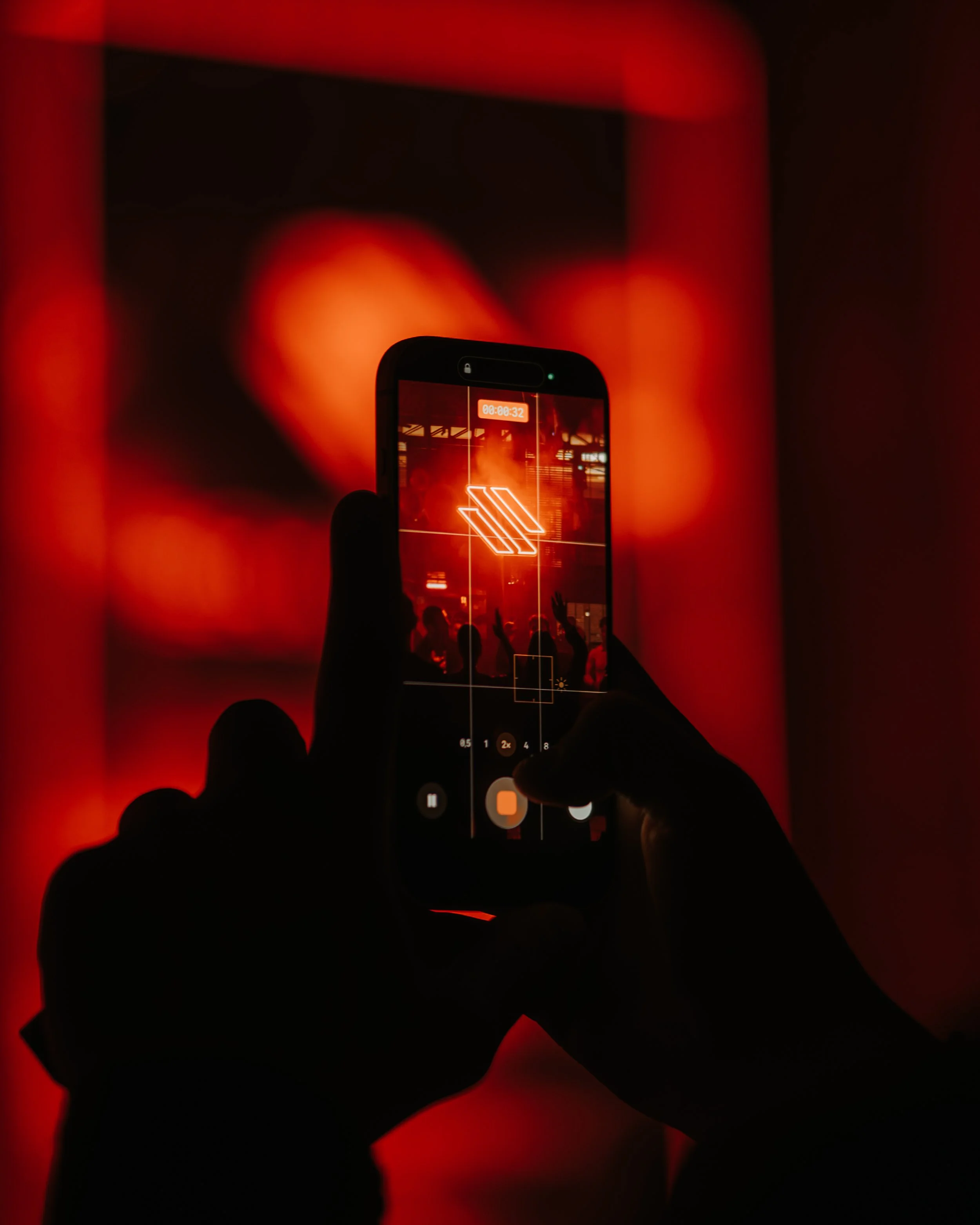 A person taking a photo or video of an event on a smartphone, with red lighting and silhouettes of people in the background.