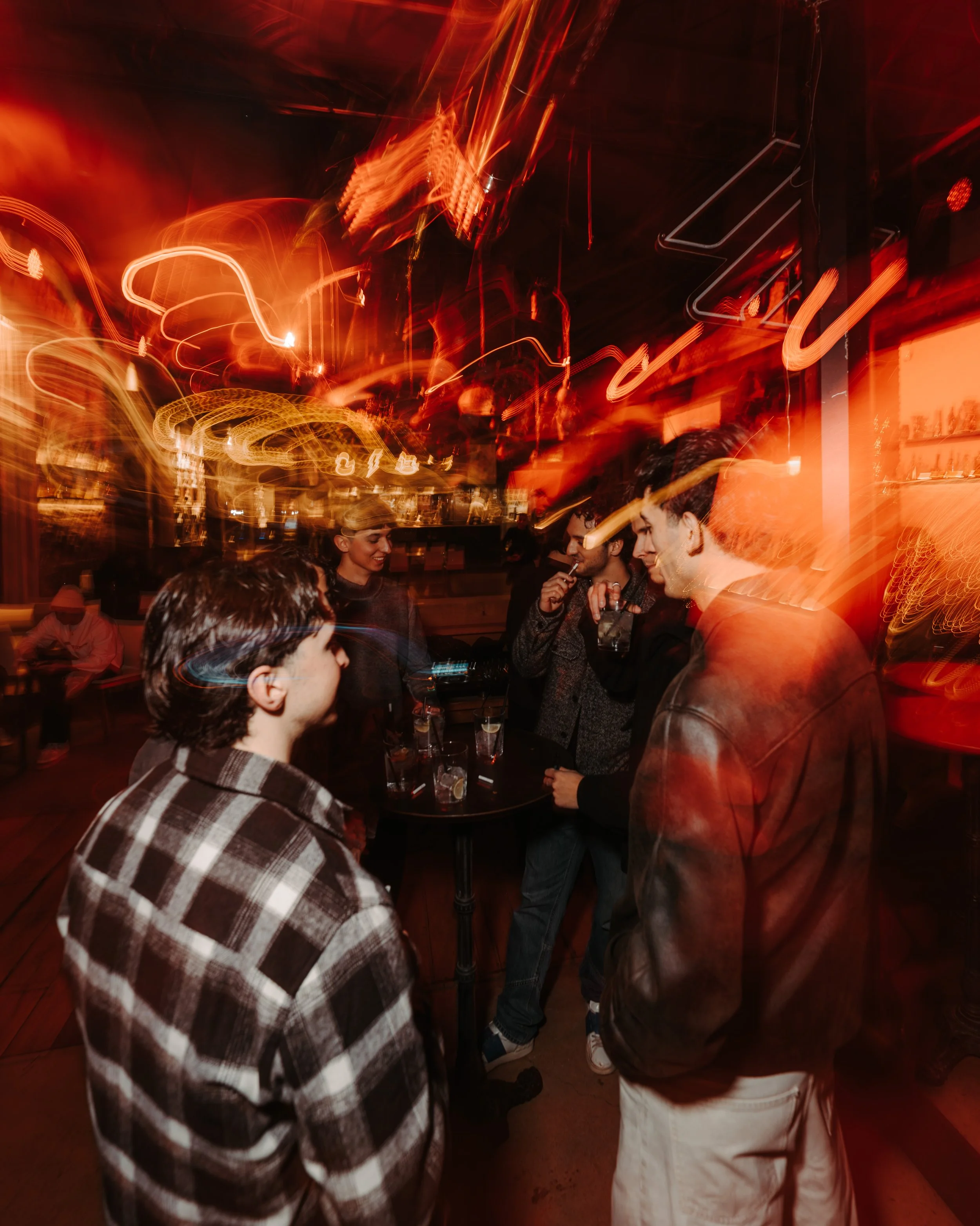 A group of young people with drinks enjoying themselves in a dimly lit bar or nightclub with red lighting and neon signs, some engaged in conversation.