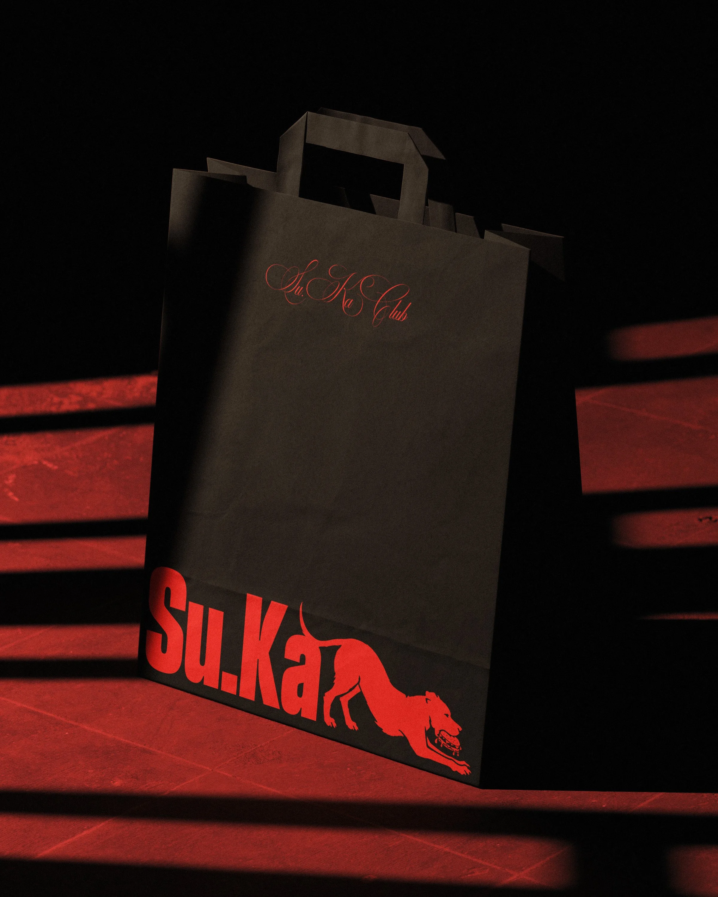 Black paper shopping bag with red and black design, featuring a running dog graphic and the text 'SU.KA CLUB',