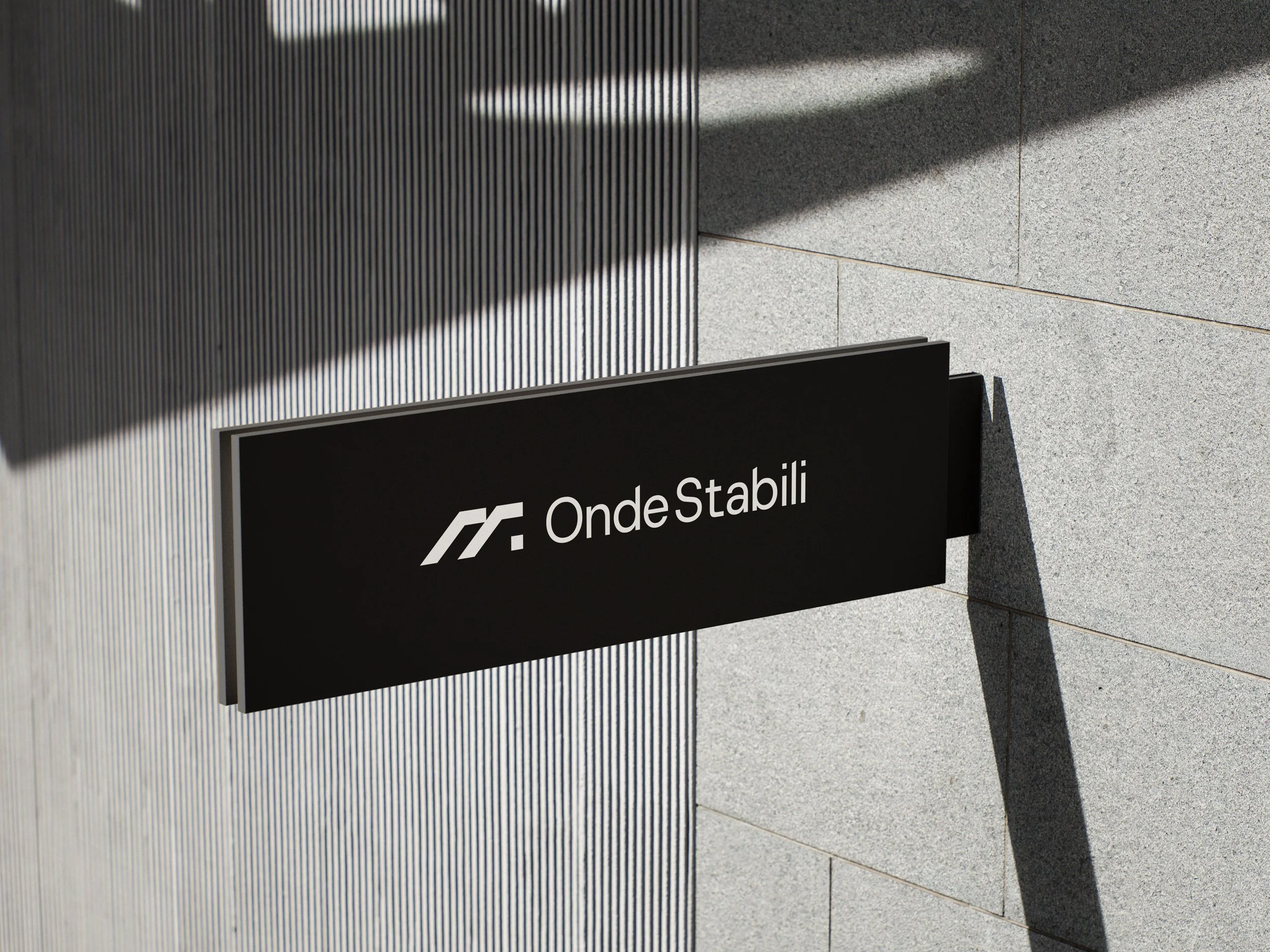 Close-up of a modern sign with the text 'Onde Stabili' and a logo, mounted on a textured gray wall with sunlight casting shadows.