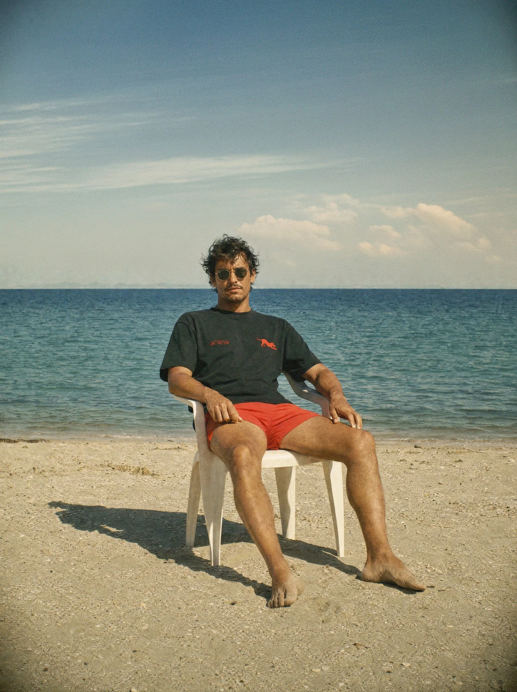 A man is sitting outdoors on a white plastic chair at the beach, wearing sunglasses, a black Su.Ka Club t-shirt, and red shorts, with the ocean and sky in the background.