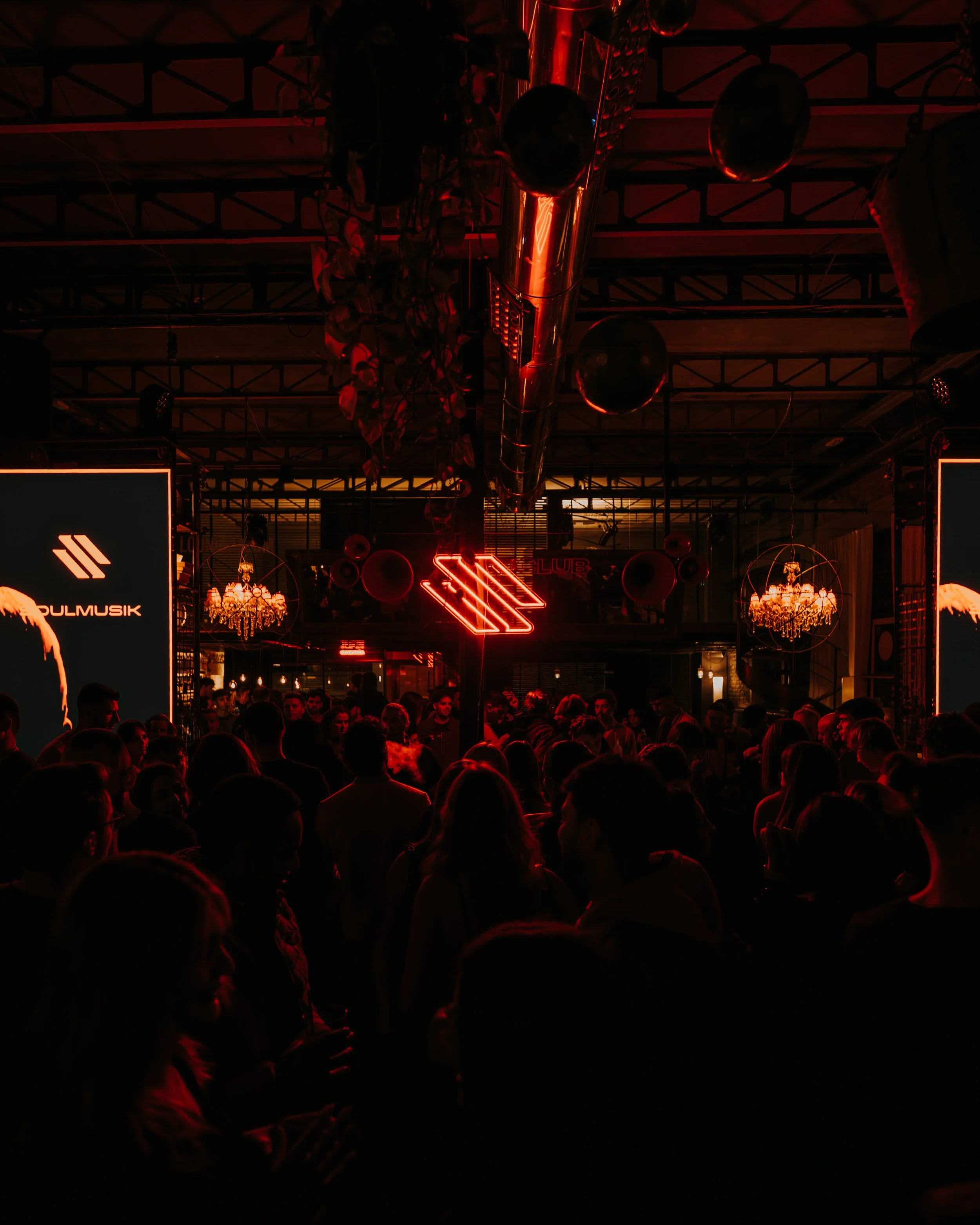 A dark interior scene with red neon lighting, including a neon logo, black balloons, chandeliers, and a crowd of people.