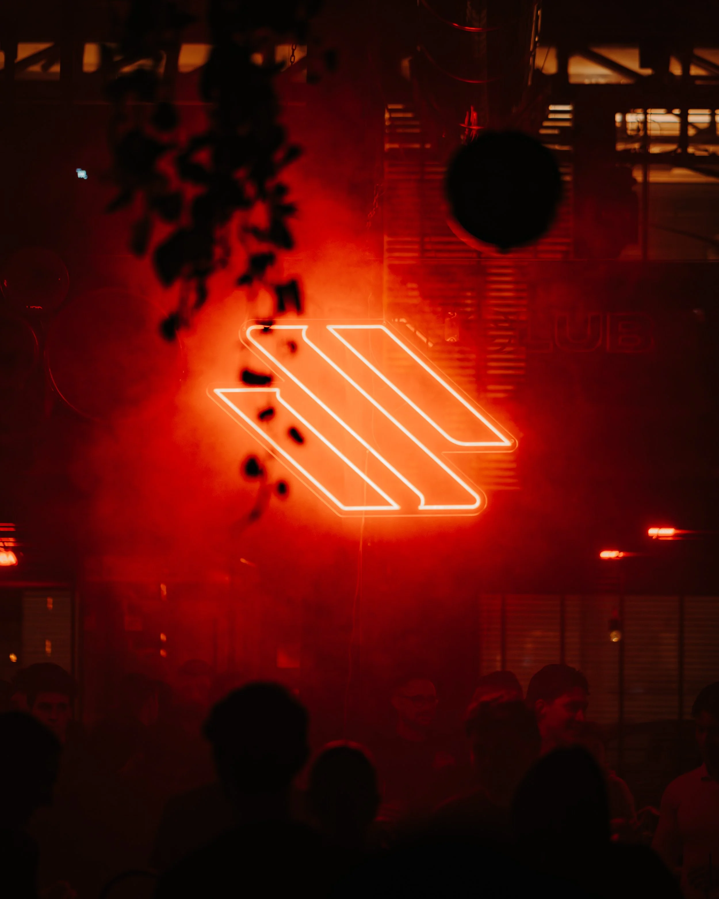 Dimly lit scene in a dark room with red lighting, featuring silhouettes of people in the audience and a stage with neon signage and a large spherical object hanging in the center.
