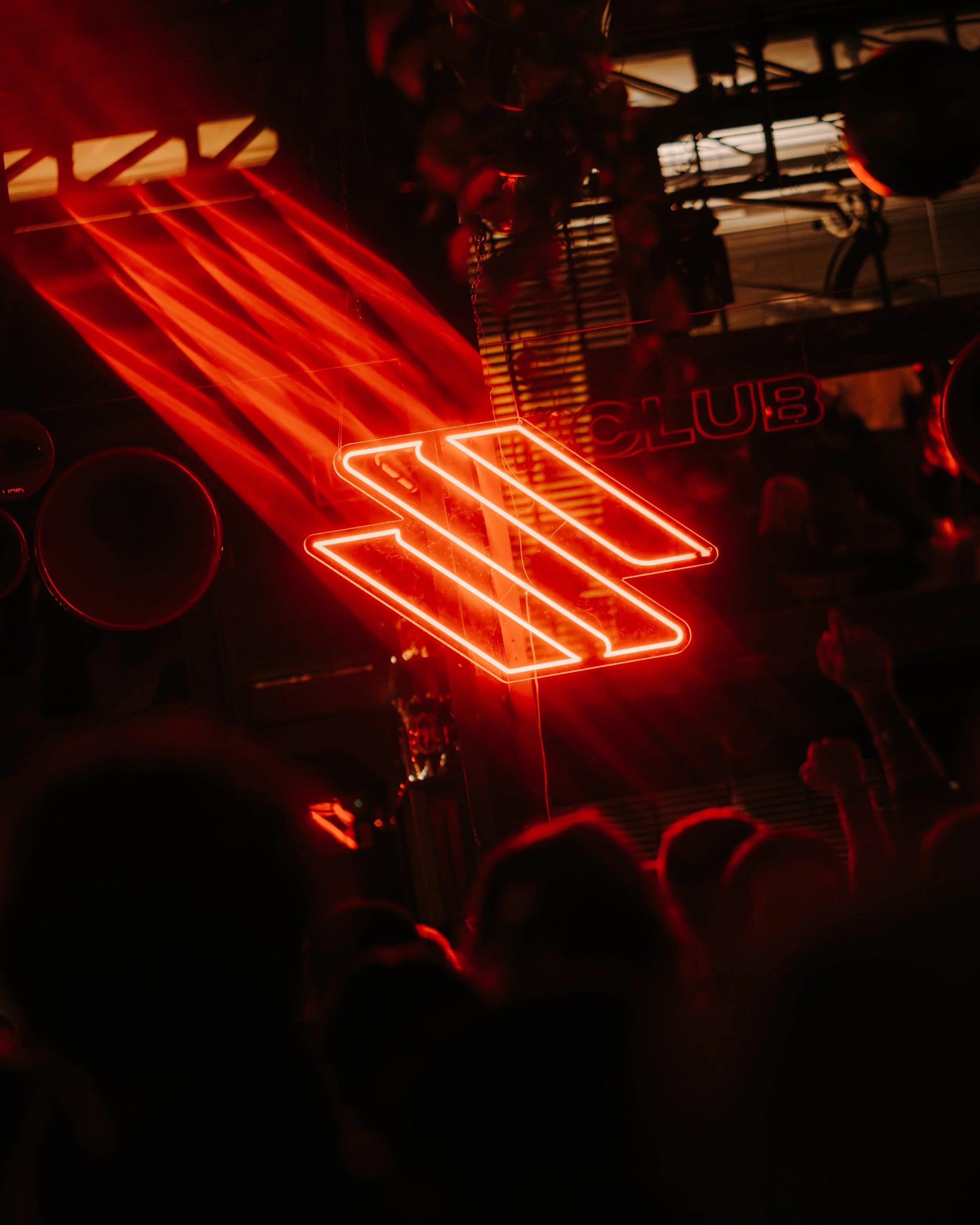 Neon sign reading 'CLUB' in red, with a large stylized arrow below it, in a dark environment with silhouettes of people.