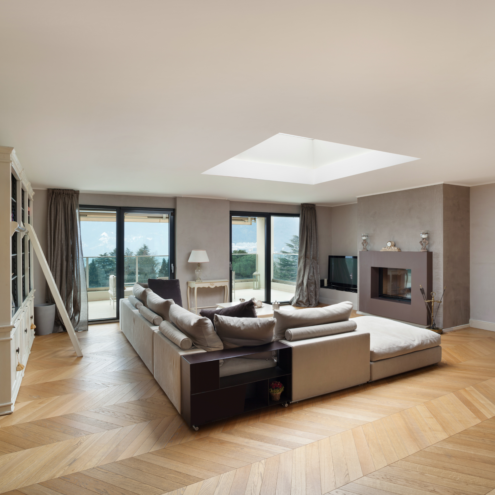 Living room with beige sectional sofa, black side table, white armchair, ceiling skylight, large windows with curtains, wooden floor, and television.