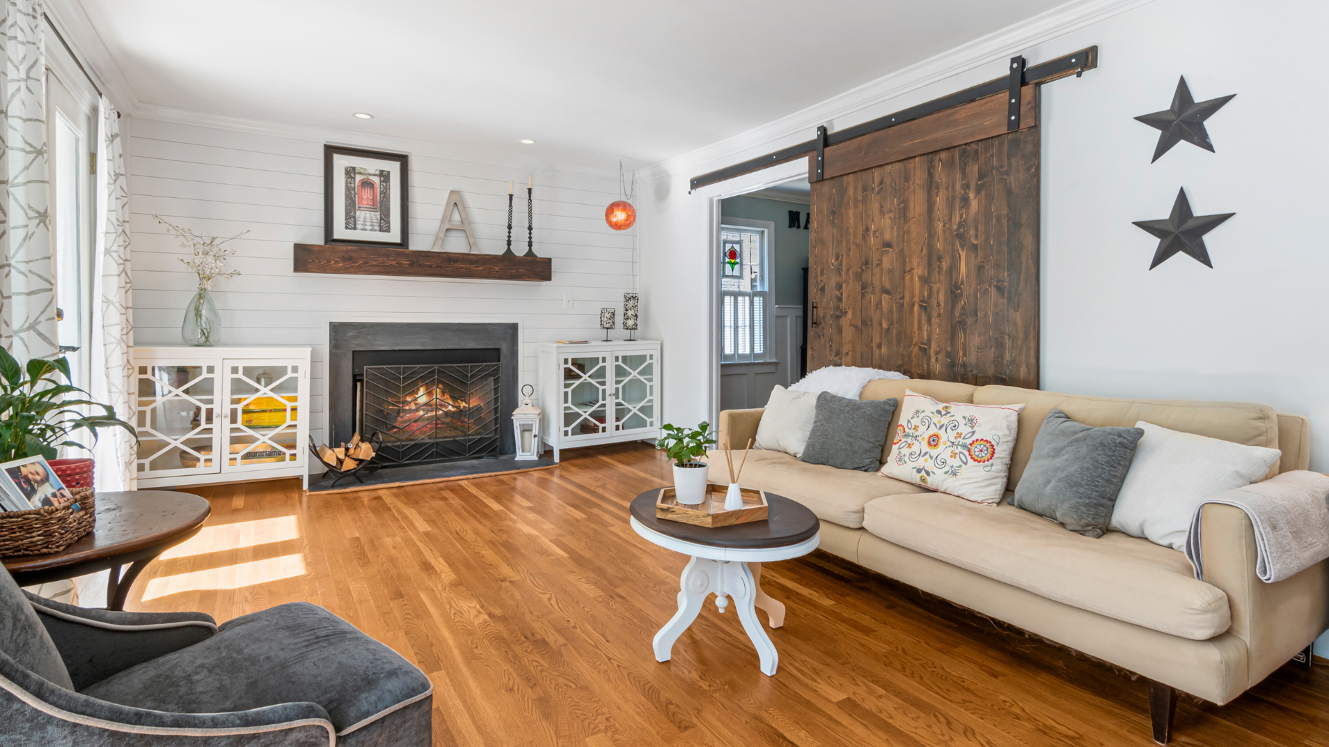 Living room with white walls, hardwood floors, a beige sofa with colorful and gray throw pillows, a dark wood sliding barn door, a fireplace with a wood mantel, and various decor items including a plant, framed pictures, star wall art, and a small co