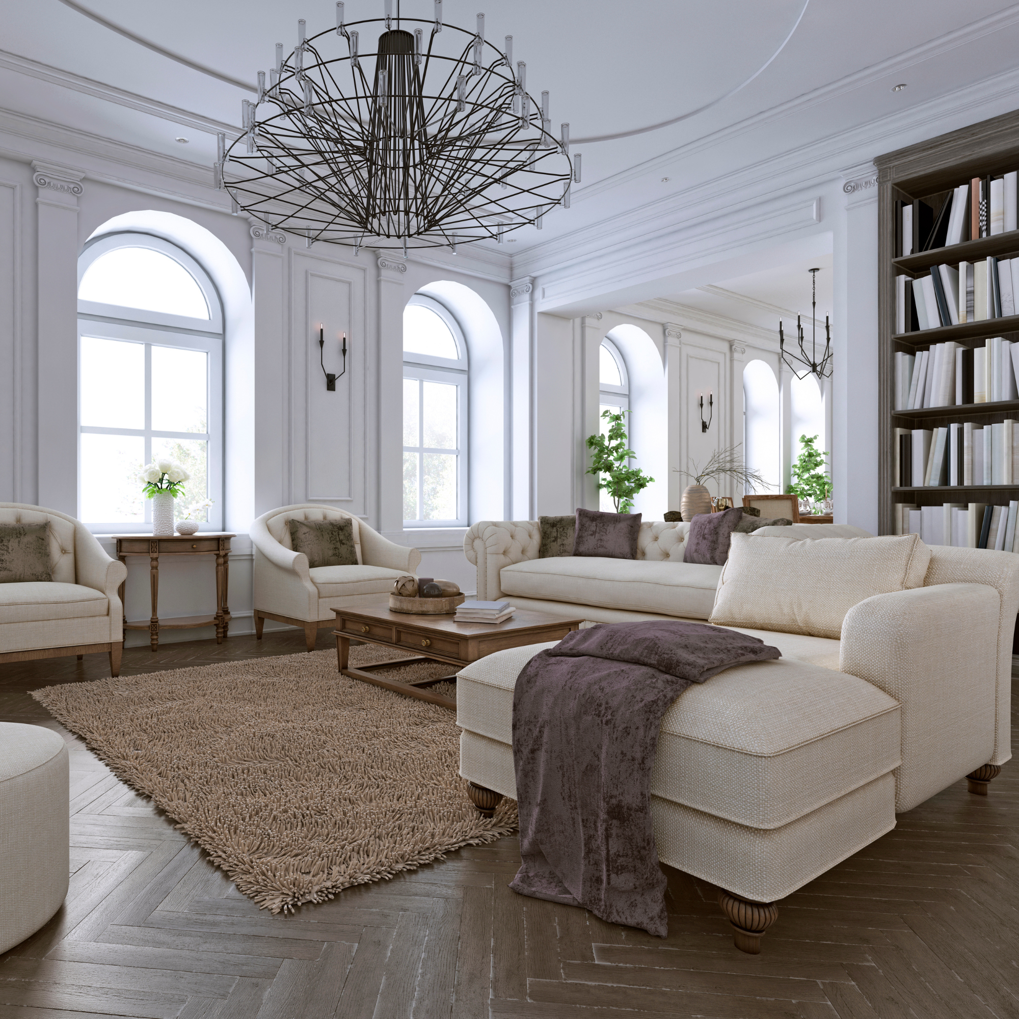 A bright living room with high ceilings, large arched windows, elegant white wall paneling, and a decorative crystal chandelier. Light-colored sofas and chairs are arranged around a wooden coffee table on a shaggy beige rug. Green plants add freshness, and a tall wooden bookshelf filled with books is partly visible on the right.