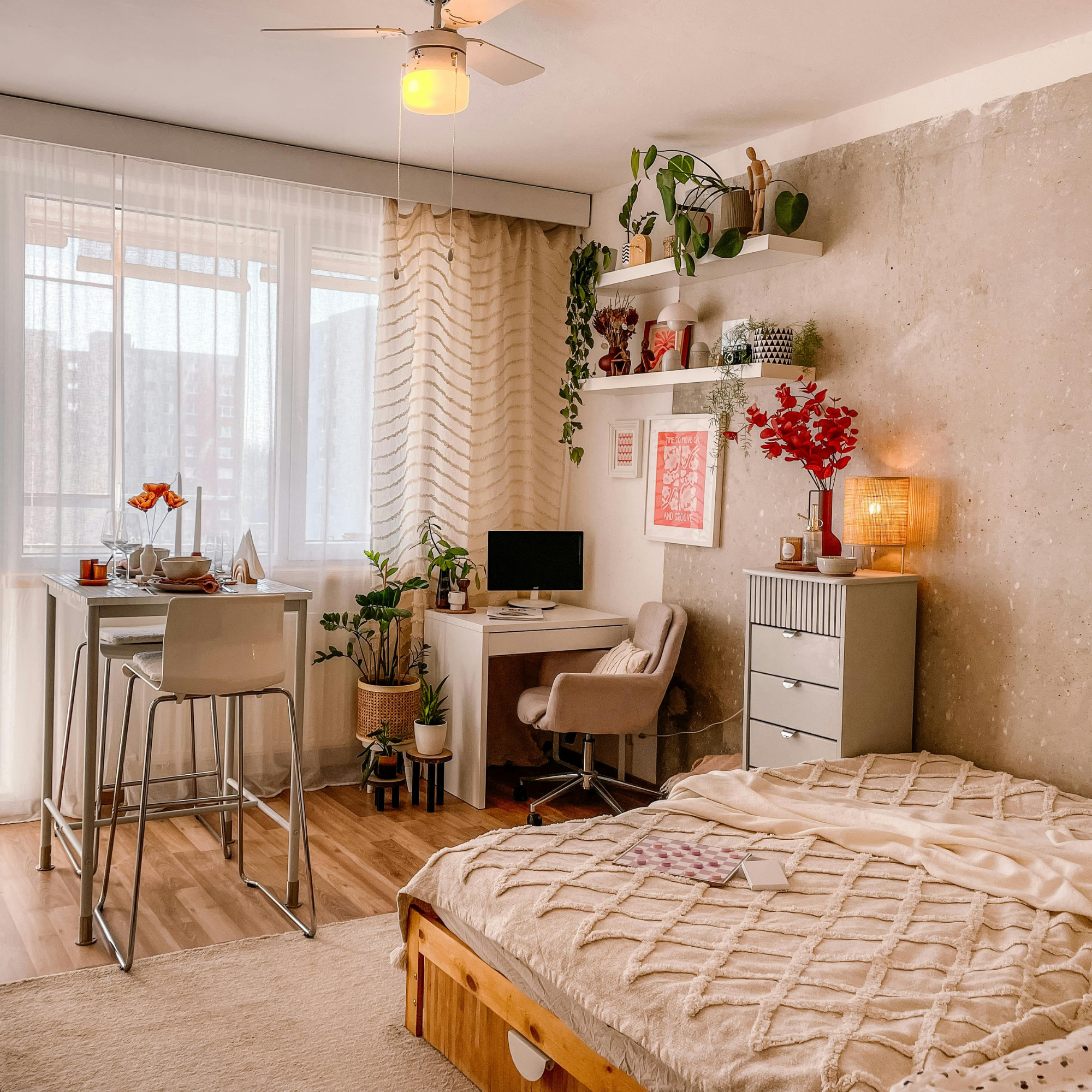 View of a cozy living space with a bed, a small dining area, and a section of a wall with floating shelves decorated with plants and art. The room has light-colored curtains, wooden flooring, and warm lighting.