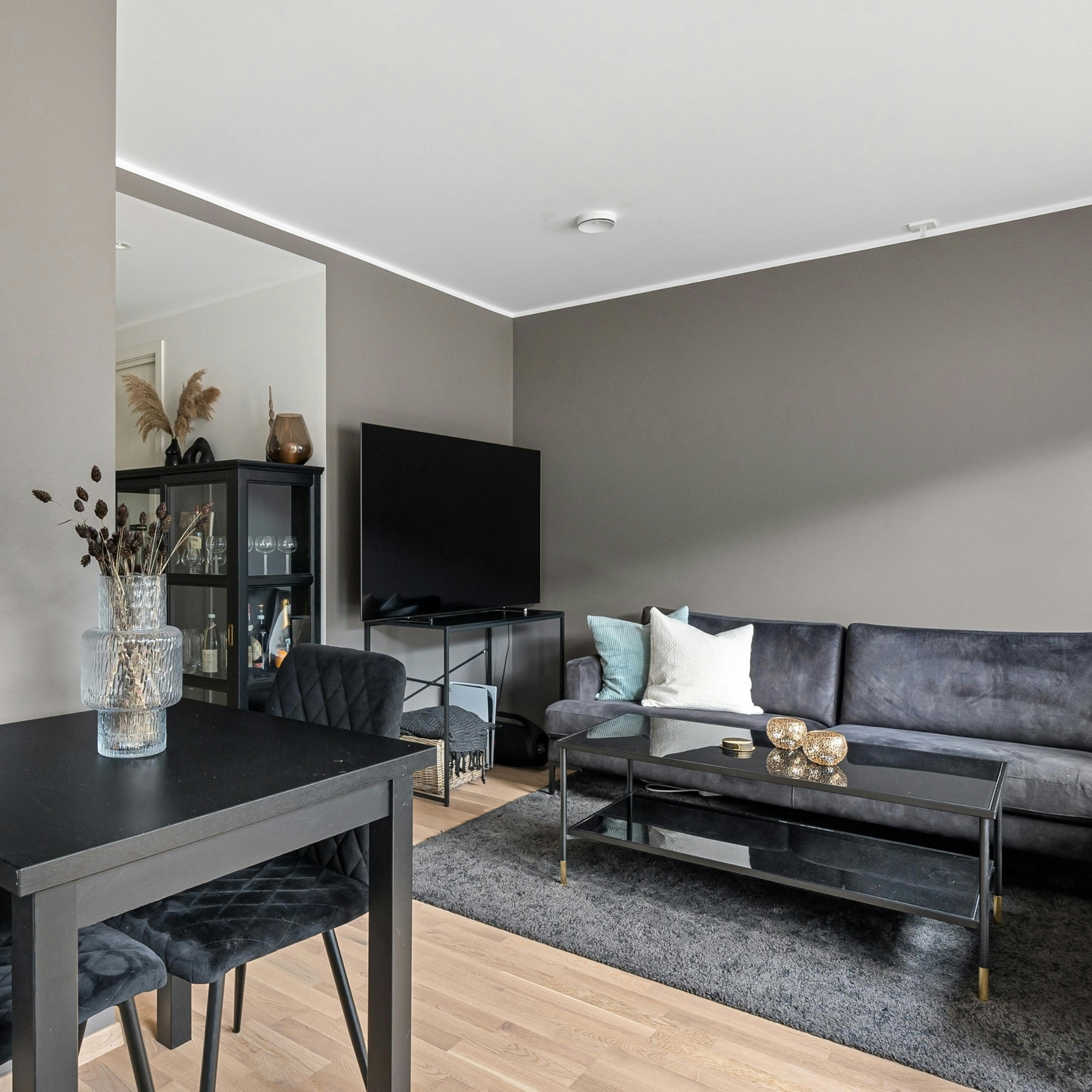 Living room with gray walls, black furniture, a flat screen TV, a black cabinet with glassware, a gray sofa with white pillows, a coffee table with gold decorative objects, a rug, and a vase with dried plants.