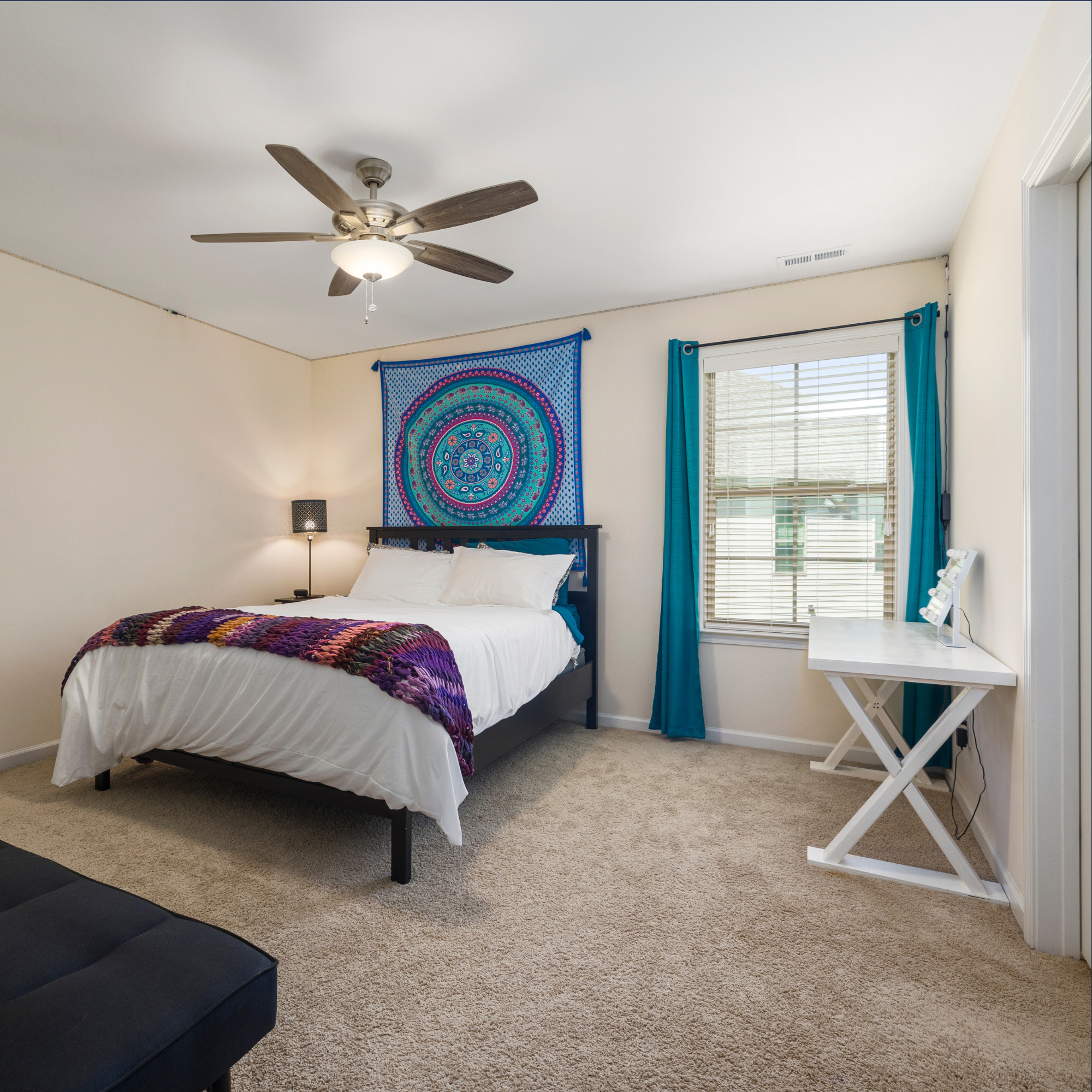 A bedroom with a white bed, colorful blanket, and a tapestry with a mandala pattern hanging on the wall. There is a nightstand with a lamp, a window with blinds and turquoise curtains, a white desk, and a ceiling fan with four wooden blades and a light fixture.