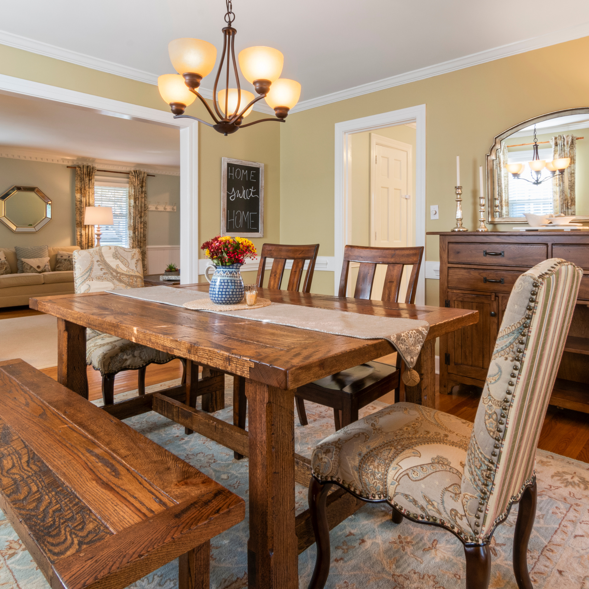 Dining room with hanging chandelier, wooden table, floral centerpiece, patterned upholstered chairs, and a mirror on the wall.
