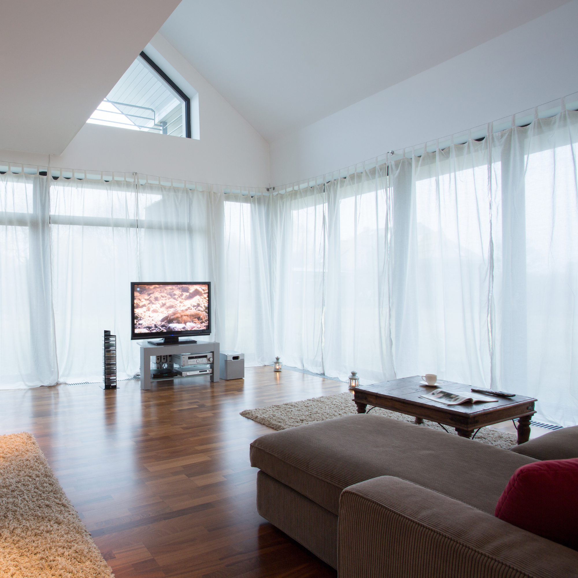 A living room with wooden floors, a brown striped sofa with red cushions, white sheer curtains, a flat-screen TV on a white stand, and small lanterns on the floor. A small table with an open magazine and cups is in the foreground.