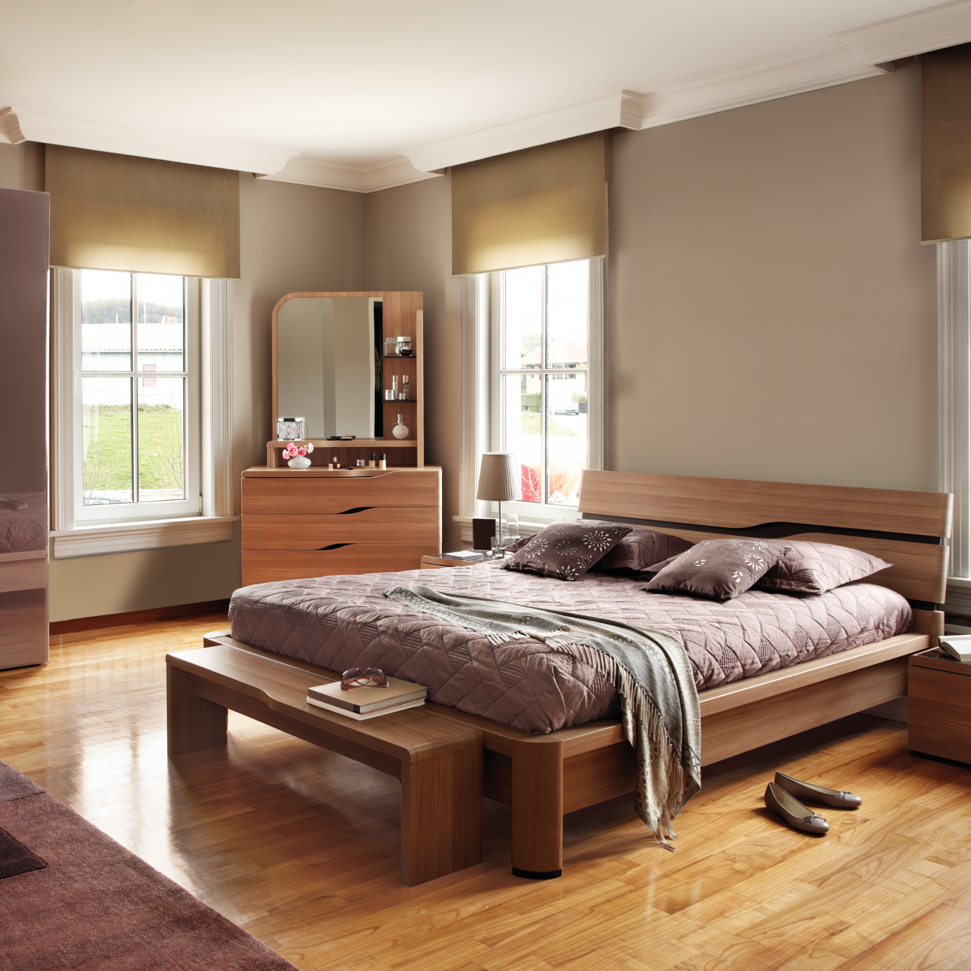 A bedroom with a wooden bed frame, two beds with brown bedding and pillows, a nightstand with shoes, a mirror, and a small shelving unit, surrounded by three large windows with beige blinds, a floor lamp, and hardwood flooring.