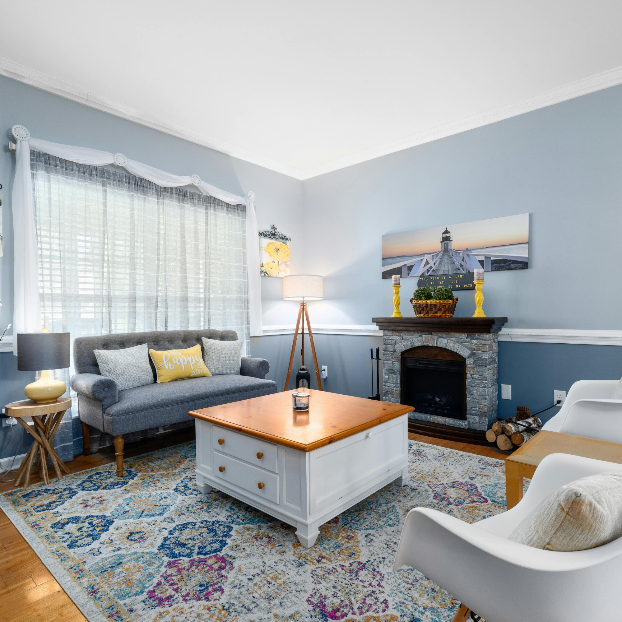 Living room with light blue walls, a fireplace with a picture of a lighthouse, a gray sofa with yellow and gray pillows, a wooden coffee table, a colorful area rug, and various decorative items including a yellow lamp, a basket of logs, and wall art.