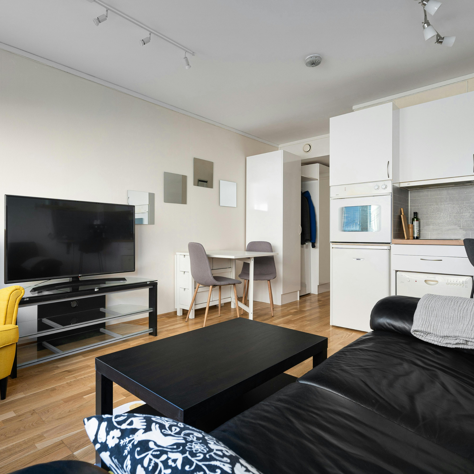 Living room with a black leather couch, wooden coffee table, yellow armchair, wall-mounted TV, and a kitchen area with white cabinets and appliances.
