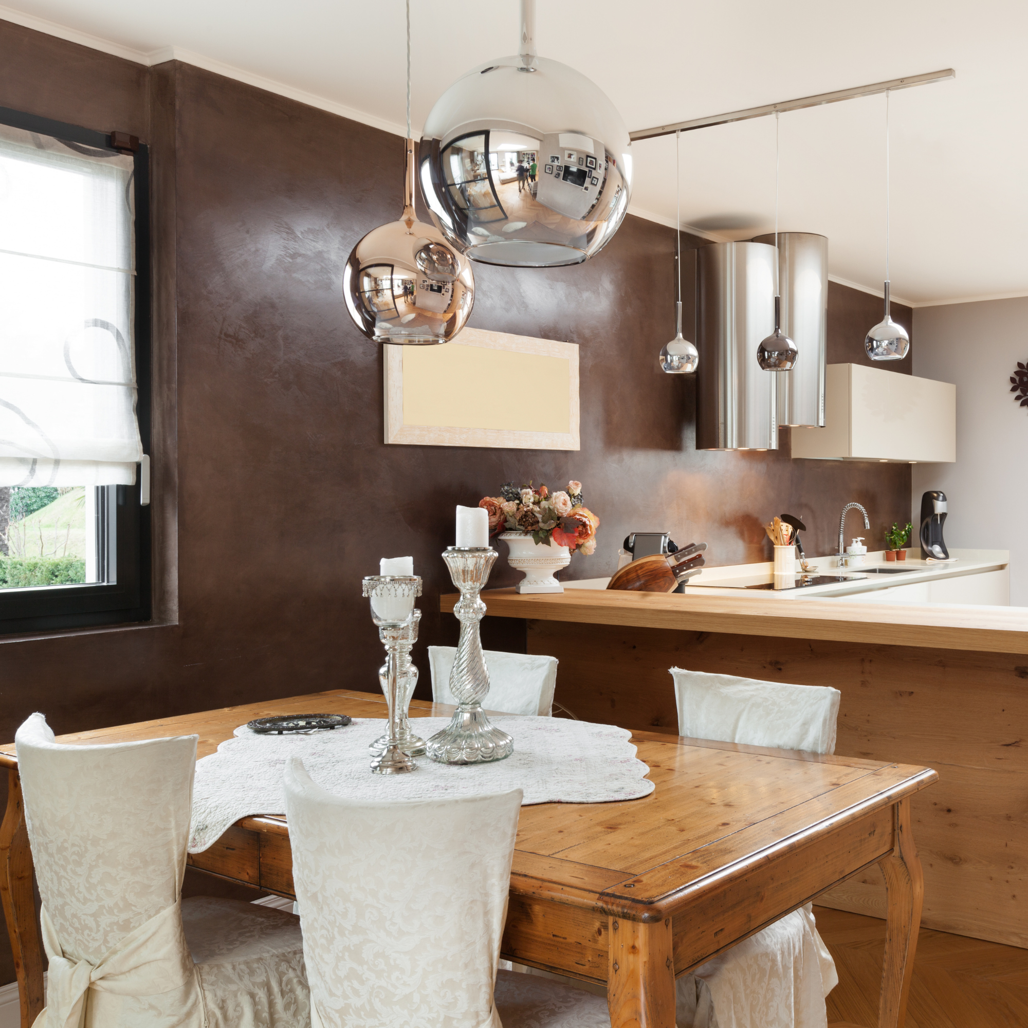 A cozy dining room with a wooden table surrounded by white cushioned chairs with slipcovers, decorated with a white lace tablecloth and a tall crystal candle holder, with a floral centerpiece. The room has a brown accent wall, a large window with whi